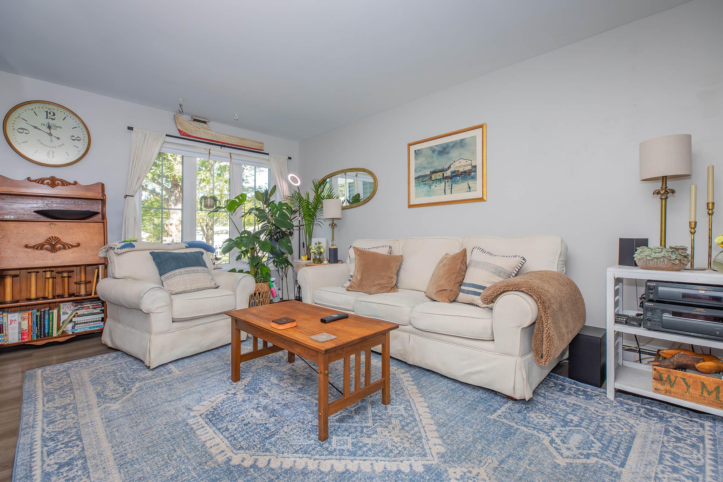A cozy living room featuring a white sofa with decorative pillows, a light armchair, a wooden coffee table, and a blue patterned area rug. There are large windows allowing natural light, a clock on the wall, a potted plant in the corner, and a small shelf with books and a media player.