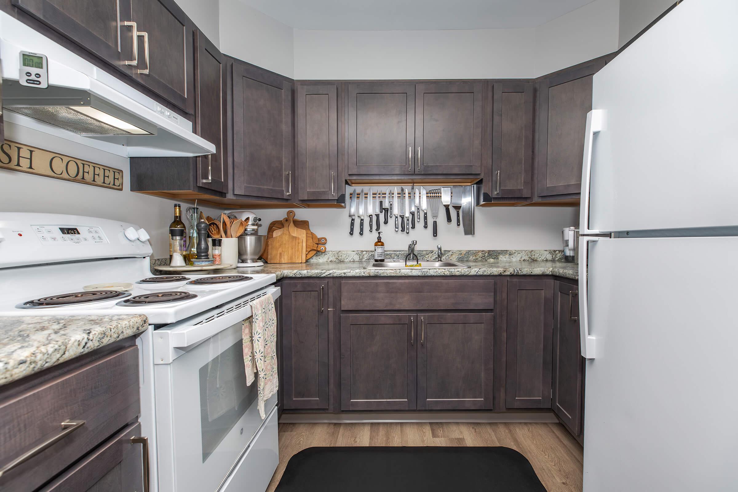 A modern kitchen featuring dark wooden cabinets, a white stove and refrigerator, a granite countertop, and various kitchen utensils hanging on a rack. The space is organized with a coffee sign displayed and a black mat on the floor, creating a cozy and functional atmosphere.