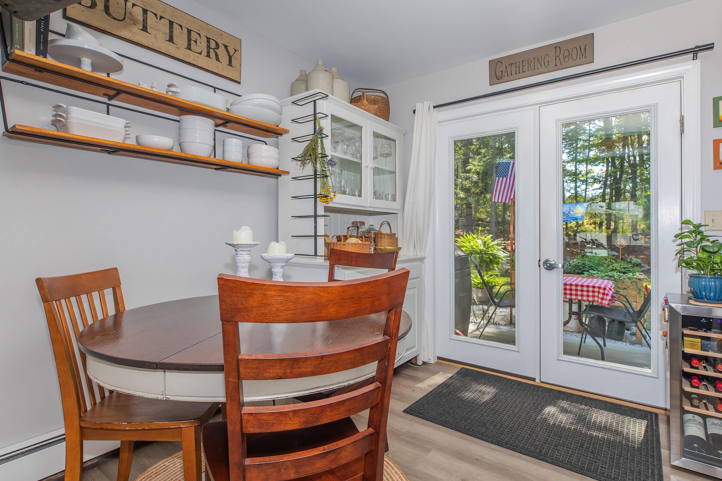 A cozy kitchen nook featuring a round wooden dining table with matching chairs, shelves displaying white dishware, and a rustic sign labeled "Gathering Room." Large glass doors lead to a patio with greenery and an American flag, enhancing the inviting atmosphere.