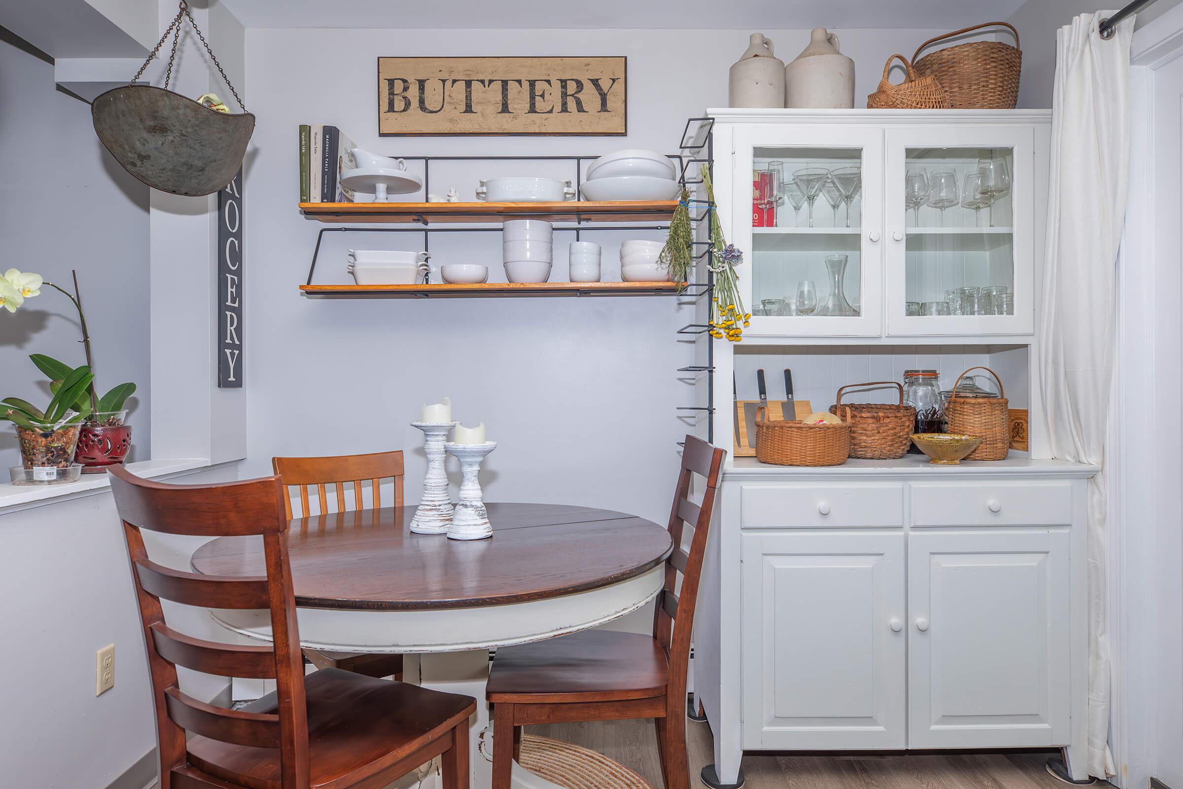 A cozy kitchen corner featuring a round wooden table with white candles on a pedestal. Surrounding it are wooden chairs. Shelves above display various kitchenware, including jars and decorative items. A white cabinet is positioned nearby, with baskets and additional kitchen essentials. A plant adds a touch of greenery.
