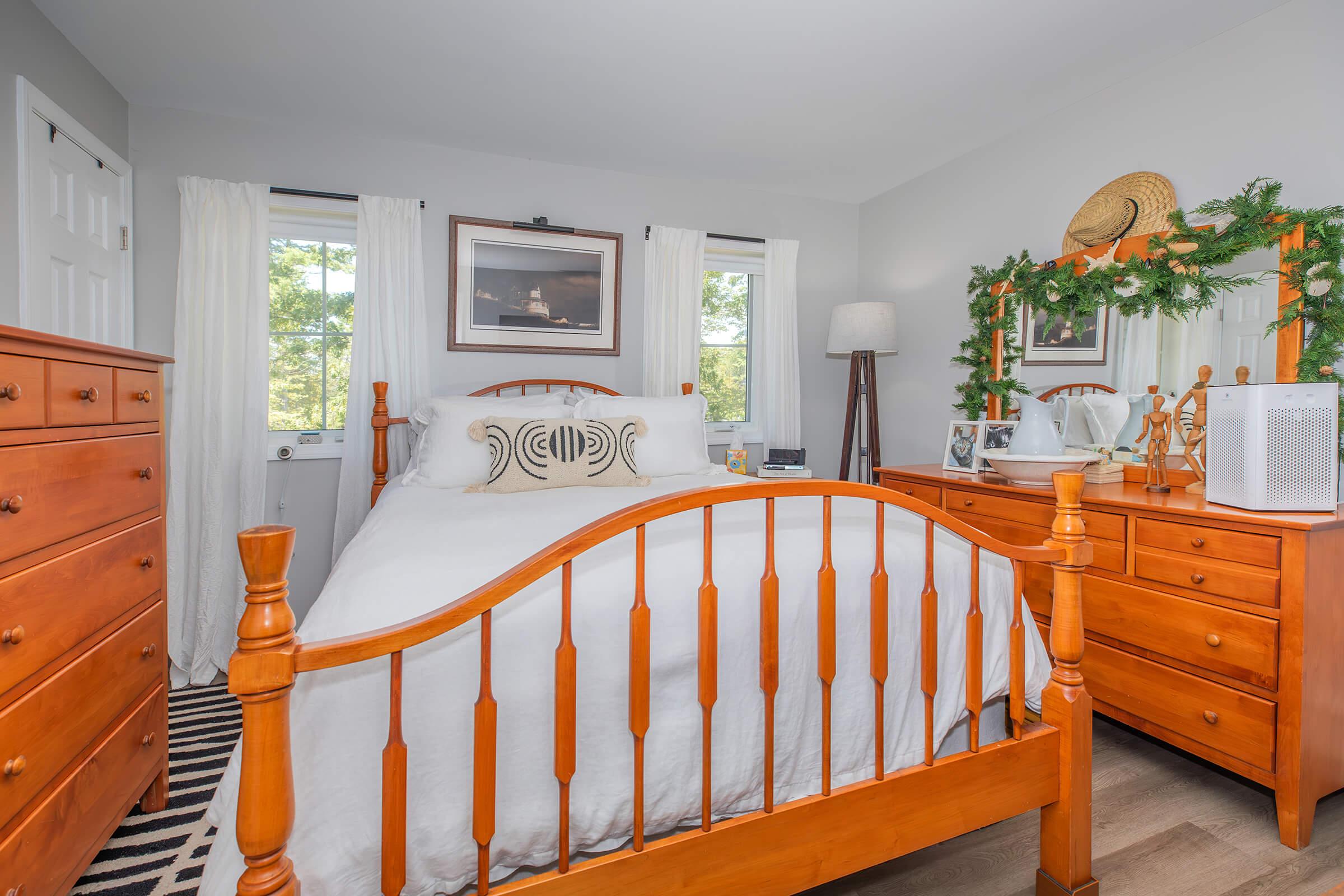 A cozy bedroom featuring a wooden bed frame with white bedding, two windows dressed with sheer curtains, and a wooden dresser. There's a lamp on a side table and decorative items on the dresser, including a framed photo and greenery. The floor has a patterned rug, creating a welcoming atmosphere.