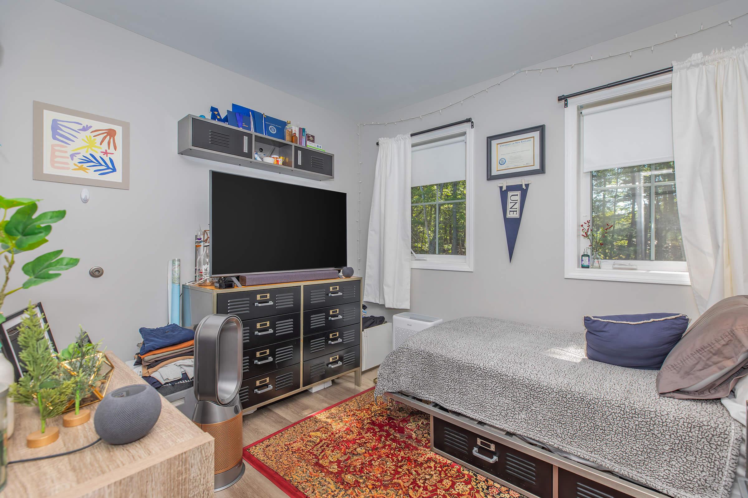 A cozy bedroom featuring a neatly made bed with gray bedding, a black media console with a large TV, and decorative items on shelves. Natural light enters through windows with white curtains. A rug adds warmth to the wooden floor, while a small plant and modern decor add a personal touch to the space.