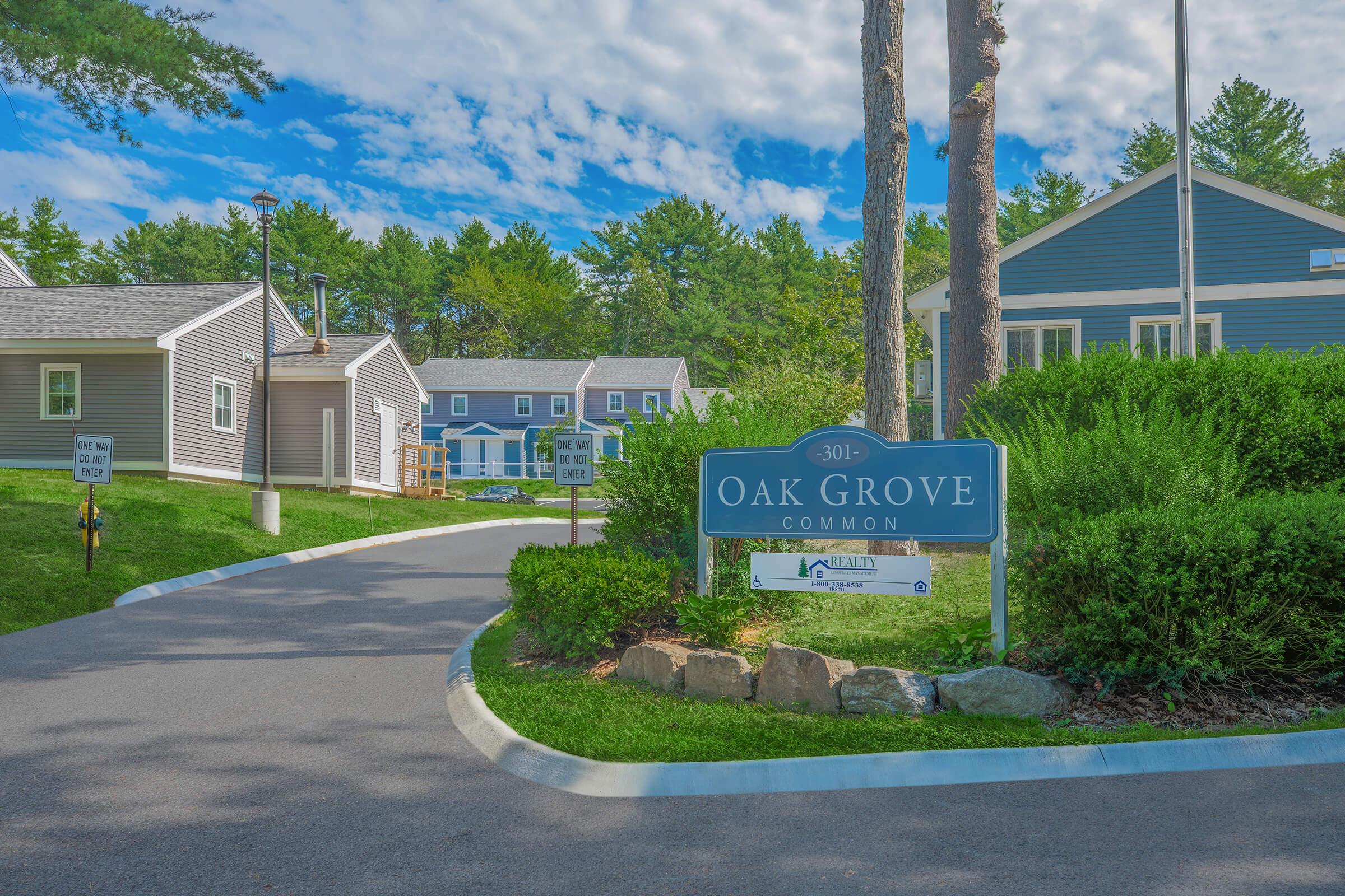 Sign reading "301 Oak Grove Common" at a residential complex entrance, surrounded by green grass and trees, with multiple houses in the background. The sky is partly cloudy, creating a serene atmosphere.