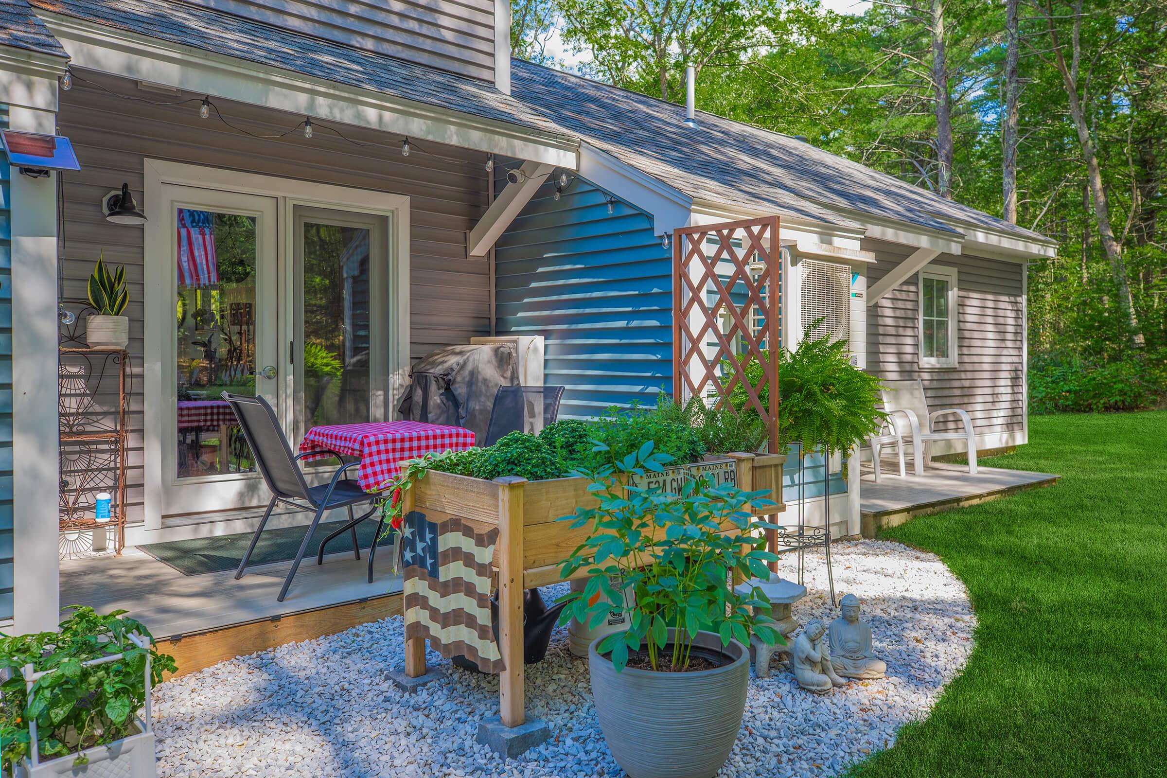 A cozy, well-maintained house with a mix of gray and blue siding, featuring a porch with seating and a checkered tablecloth. A decorative planter with greenery and a small statue are in the foreground, surrounded by white gravel and a lush lawn. Trees provide a serene backdrop.