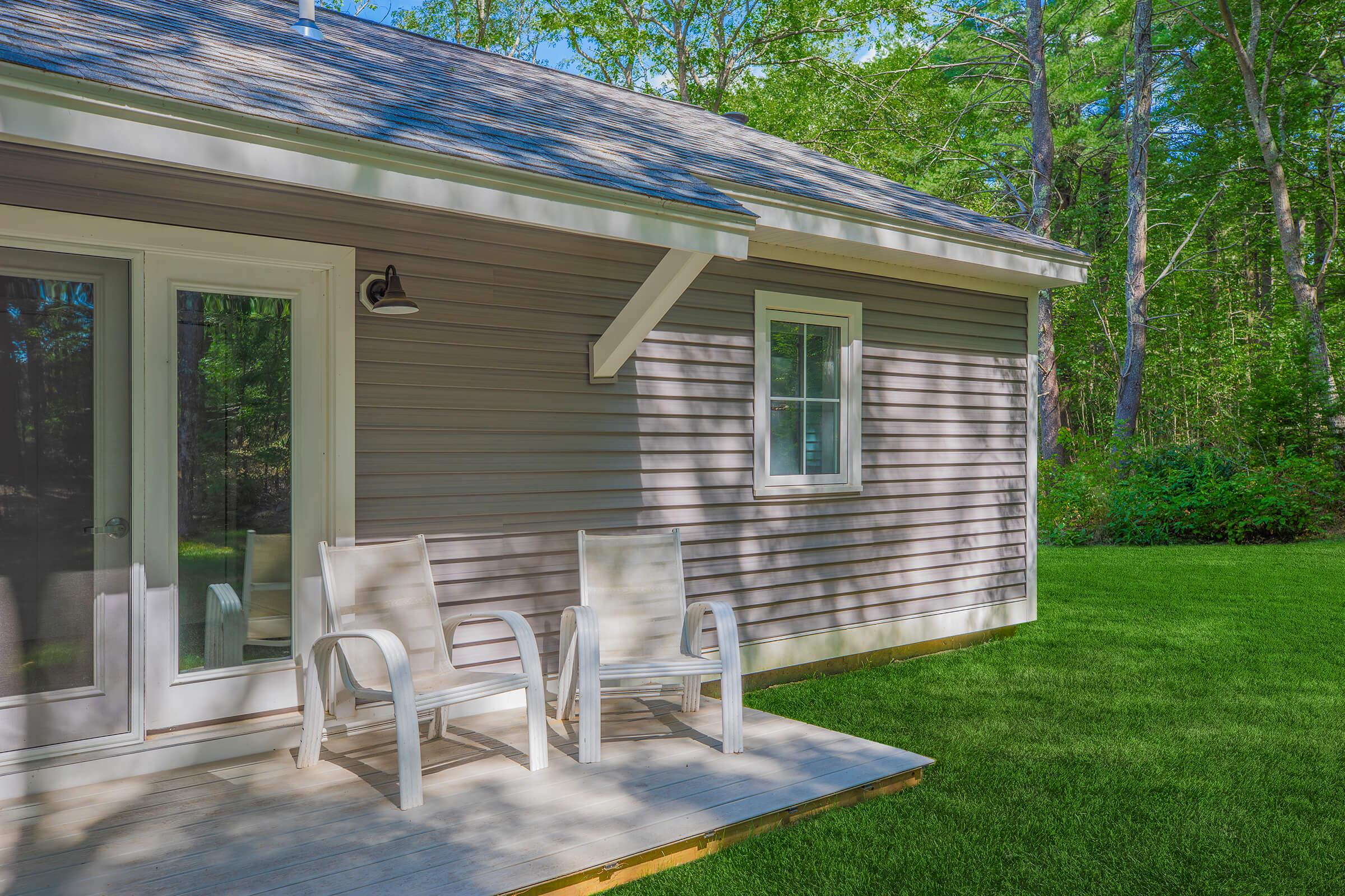 A cozy cottage with a light gray exterior, featuring a wooden deck and two white chairs. The surrounding area has vibrant green grass and lush trees, creating a peaceful, natural setting. Sunlight casts shadows on the house, enhancing its charming atmosphere.