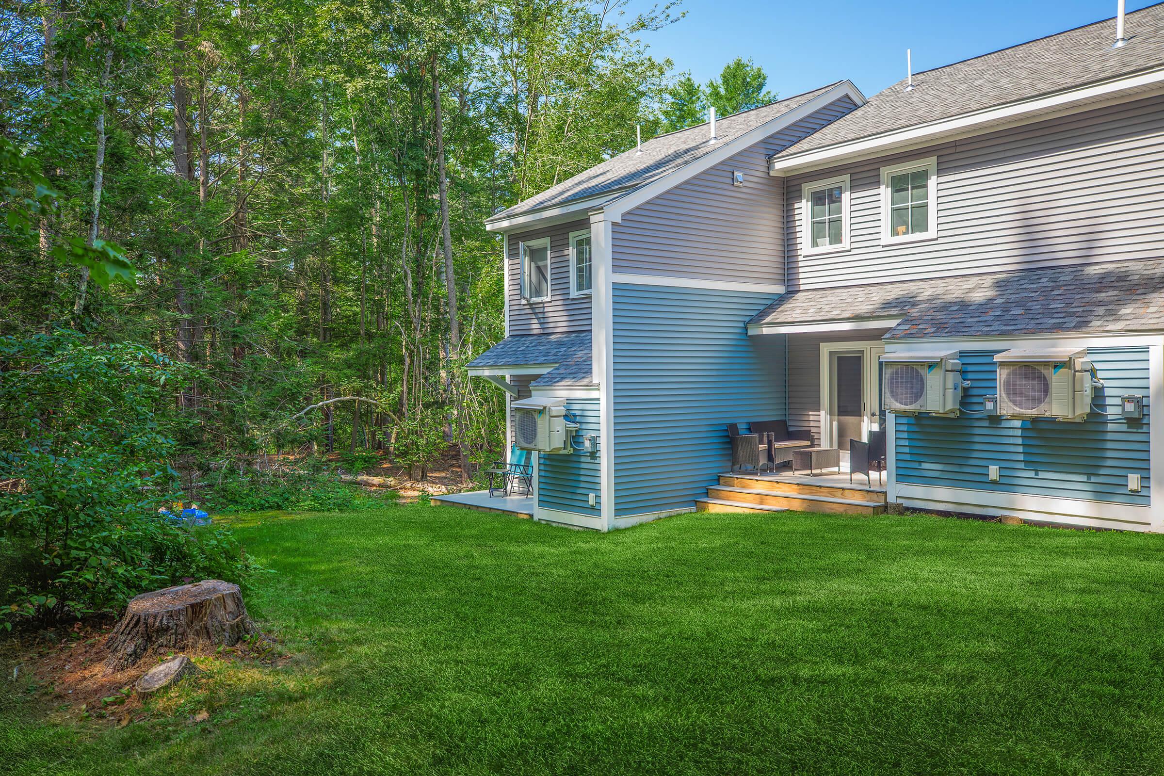 A backyard view of a two-story house featuring blue and gray siding, with air conditioning units mounted on the side. The well-kept yard has lush green grass and a tree stump, surrounded by dense trees providing a serene, natural setting.