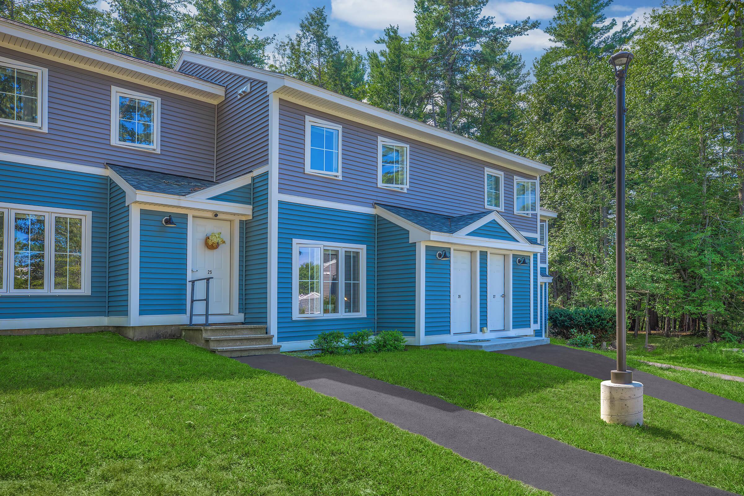 A vibrant blue multi-unit building surrounded by lush green grass and tall trees. The structure features two entrances with white doors, a small porch area, and a well-maintained pathway leading to the entrance. Bright daylight enhances the cheerful atmosphere of the scene.