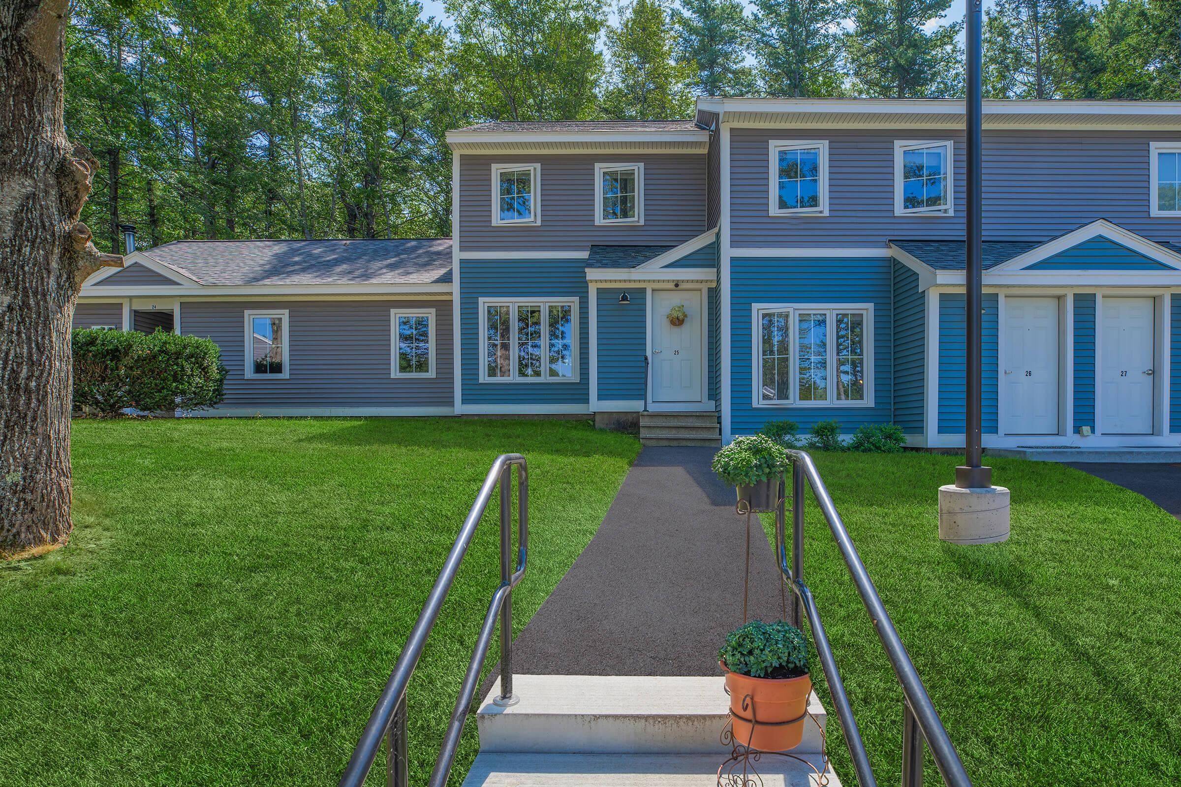 A modern two-story building with blue siding, featuring large windows and a well-maintained lawn. A concrete pathway leads to the entrance, with potted plants on the steps. Surrounding trees provide a lush backdrop, creating a welcoming outdoor environment.