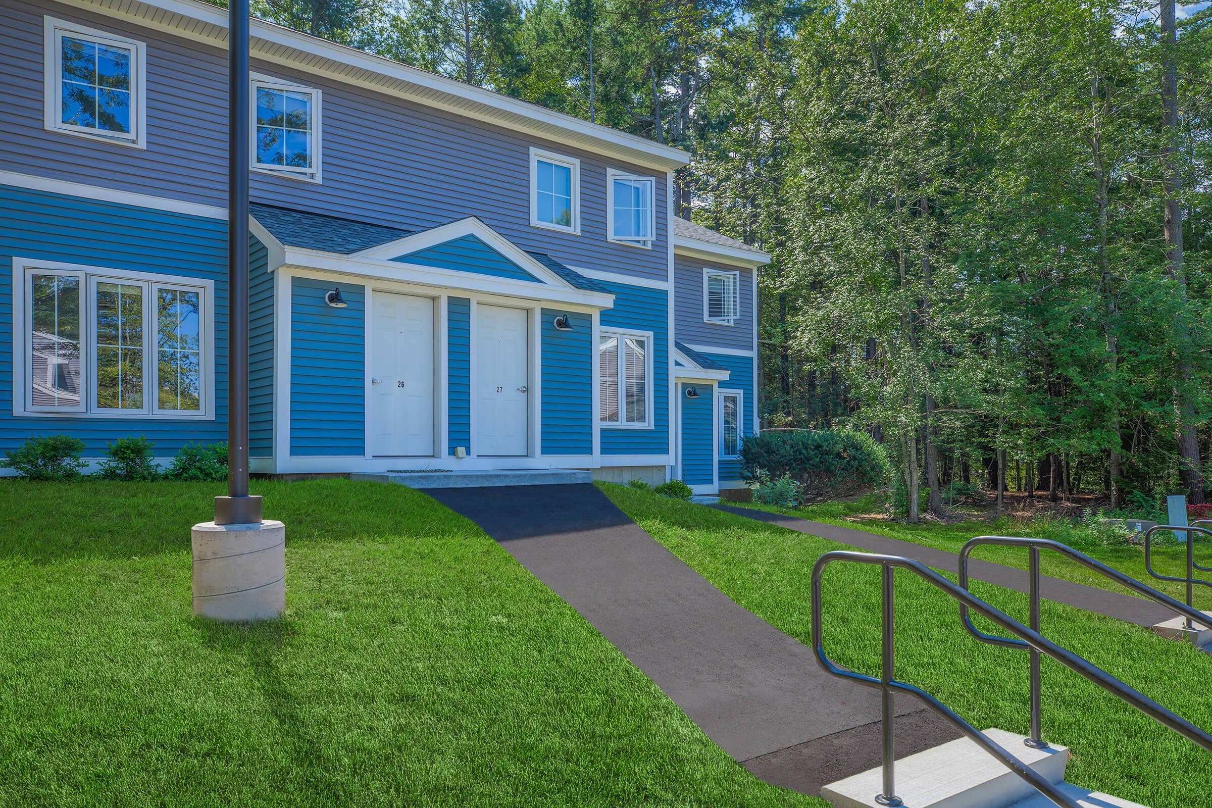 A modern two-story building with blue siding and white trim, set in a lush, green landscape. The front features two white doors, a concrete walkway, and a handrail leading to the entrance. Surrounding vegetation includes trees and manicured grass. The scene is bright and inviting.