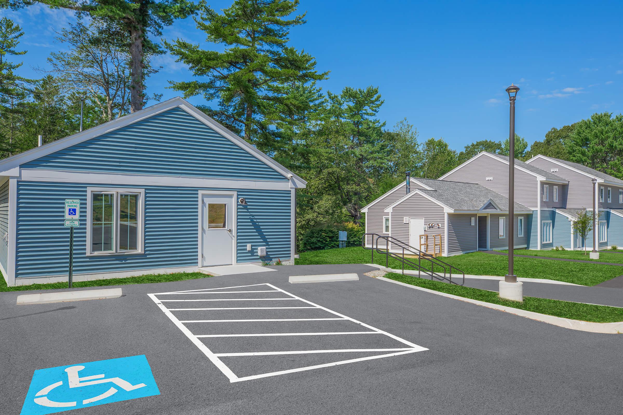 A blue handicapped-accessible parking space in front of a light blue building with a white door. In the background, a light gray building is visible, surrounded by trees and grass. The scene is set on a sunny day with a clear blue sky.
