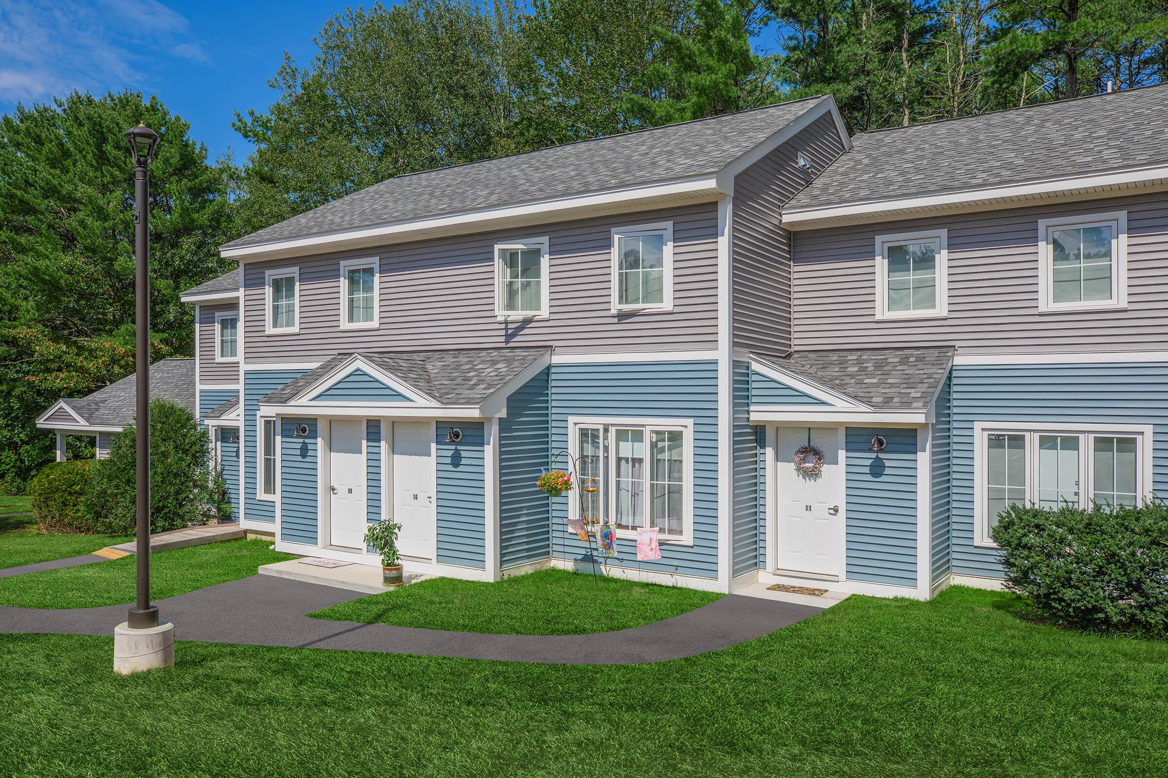 A row of two-story residential buildings, featuring a mix of blue and beige siding. The homes have front doors with small porches, windows adorned with flower pots, and well-maintained lawns. A paved walkway leads to the front doors, set against a backdrop of green trees and a clear blue sky.