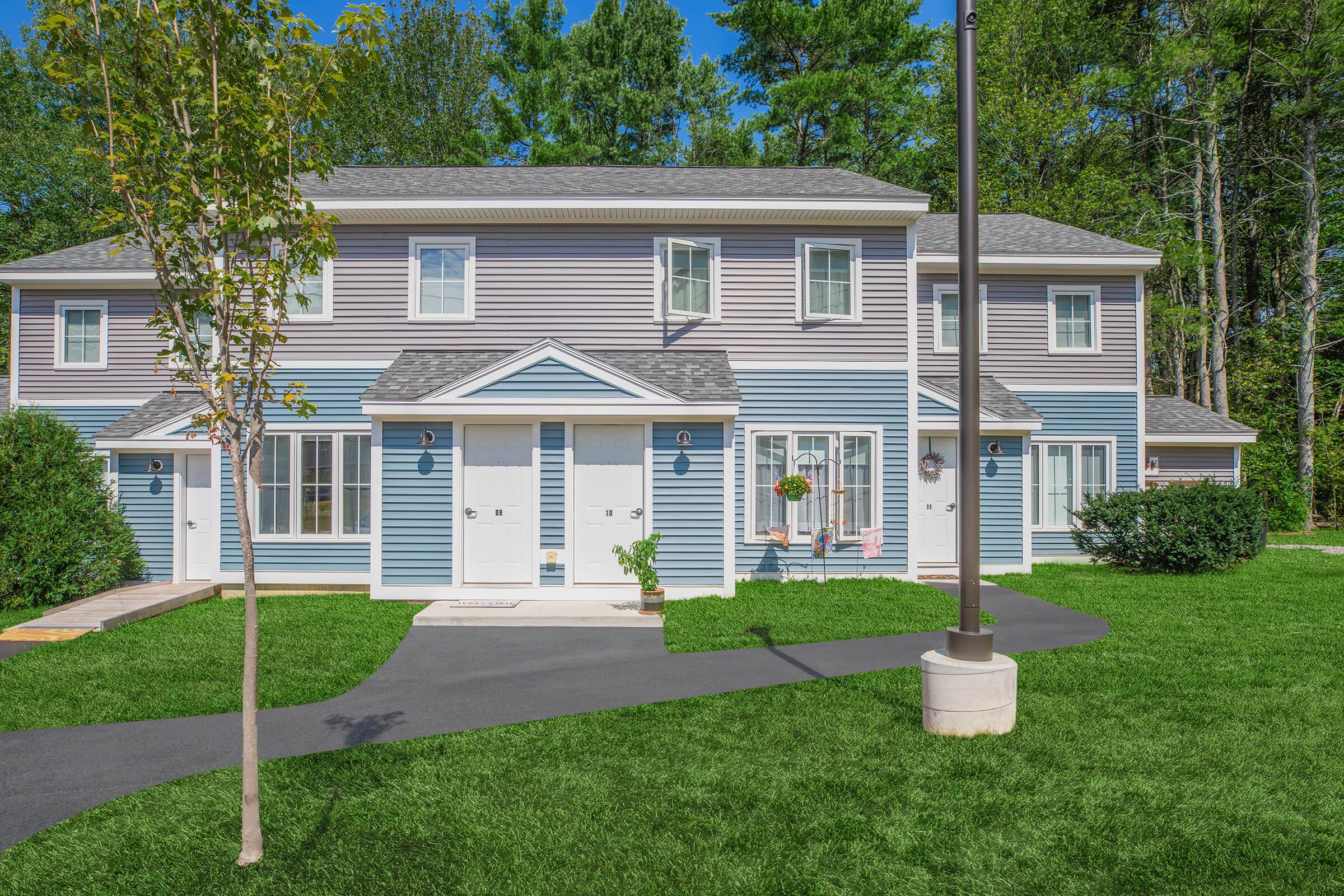 A modern two-story residential building with blue and gray exterior, featuring multiple entrance doors. The surrounding area includes green grass, small trees, and a paved walkway. Bright sunlight illuminates the scene, giving it a cheerful atmosphere.