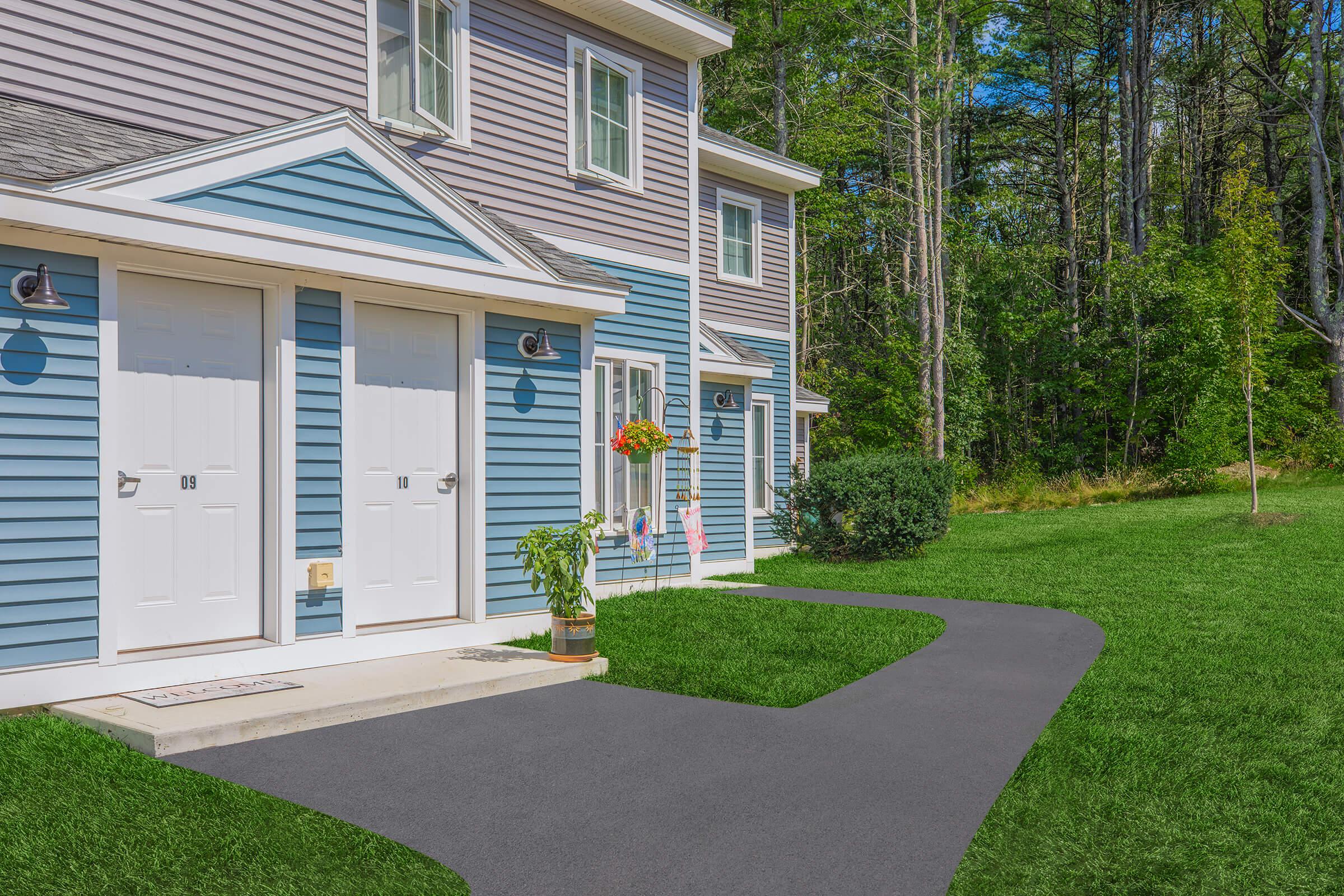 A view of a two-story building with blue and gray siding, featuring separate white doors labeled 109 and 110. A grassy pathway leads to the entrance, with a flower pot and a hanging flower basket adding a pop of color. Lush greenery and trees are visible in the background, creating a serene outdoor setting.