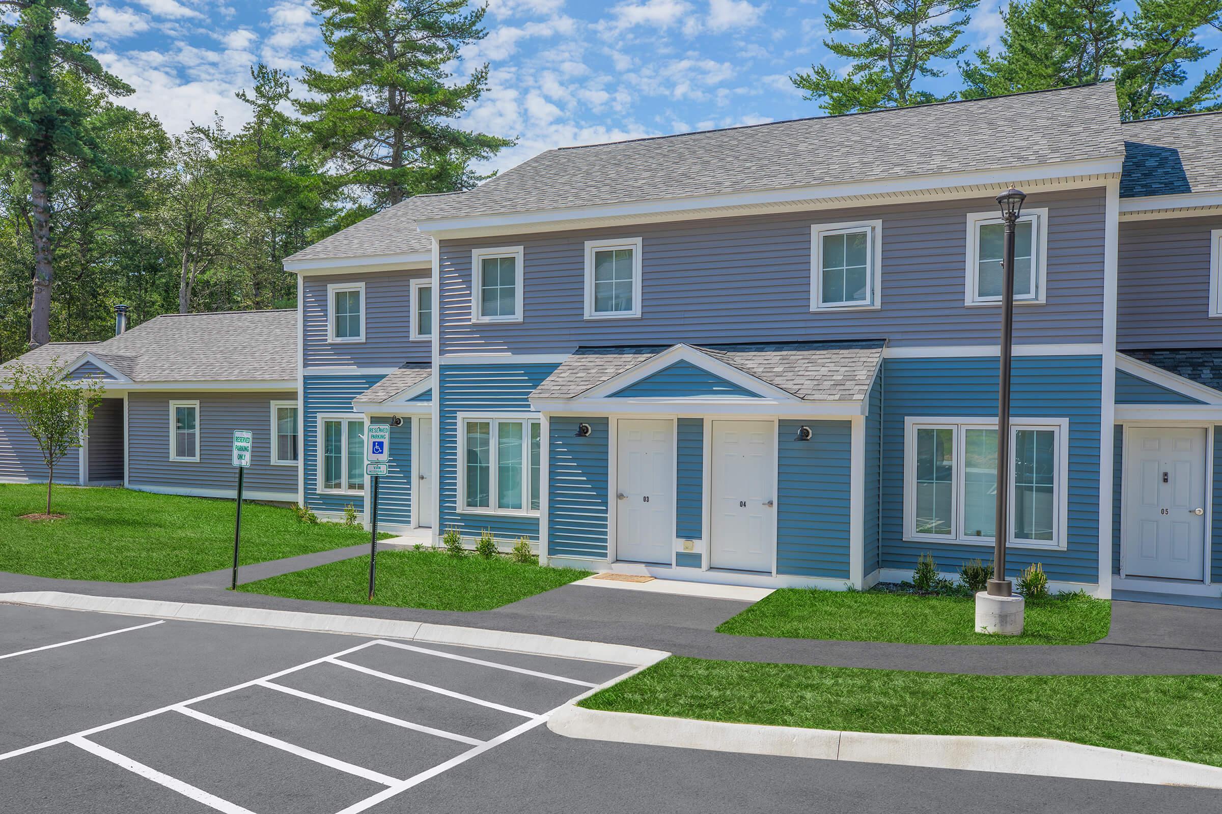 A row of modern two-story townhouses with blue siding and white trim, surrounded by green grass and trees. The houses feature porches, windows, and doors in a well-maintained setting. A parking lot is visible in the foreground, along with painted lines for parking. The sky is clear with a few clouds.