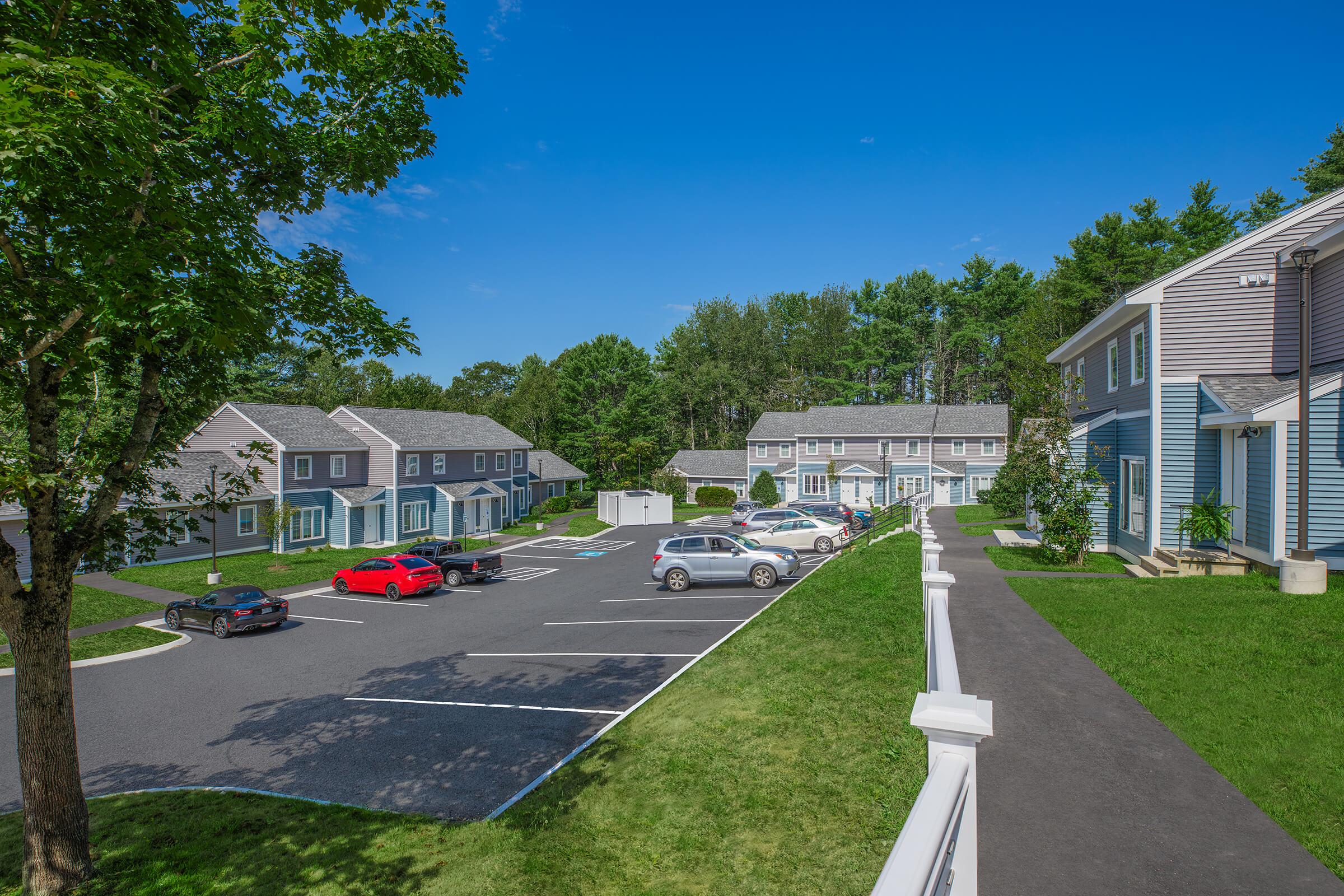 A scenic view of a landscaped residential area featuring multi-unit houses. Numerous vehicles are parked in a lot, with green trees in the background and clear blue skies above. A pathway leads through the grassy areas, enhancing the inviting atmosphere of the community.