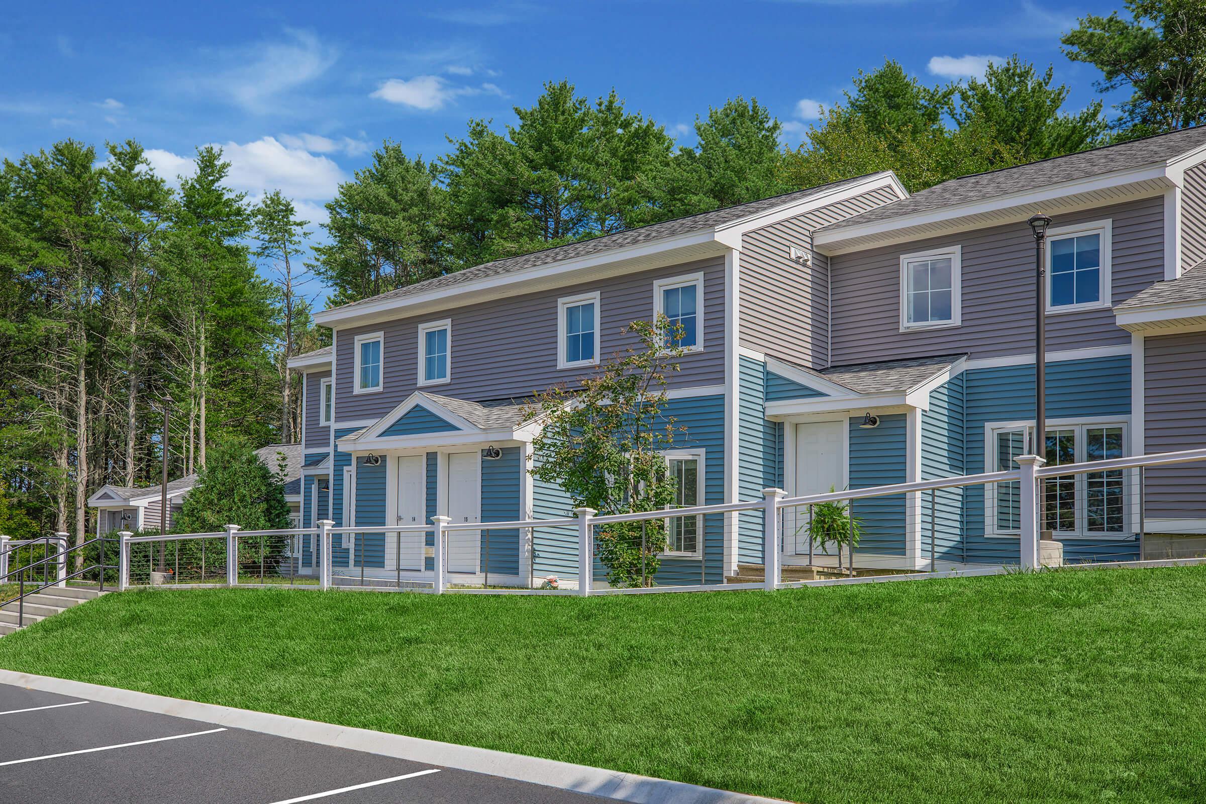 A row of modern townhouses in a residential area, featuring a mix of blue and gray siding. Each unit has a small porch and landscaping with trees in the background. A paved parking area is visible in the foreground, with neatly arranged parking spaces. The scene is bright and inviting, with clear blue skies.