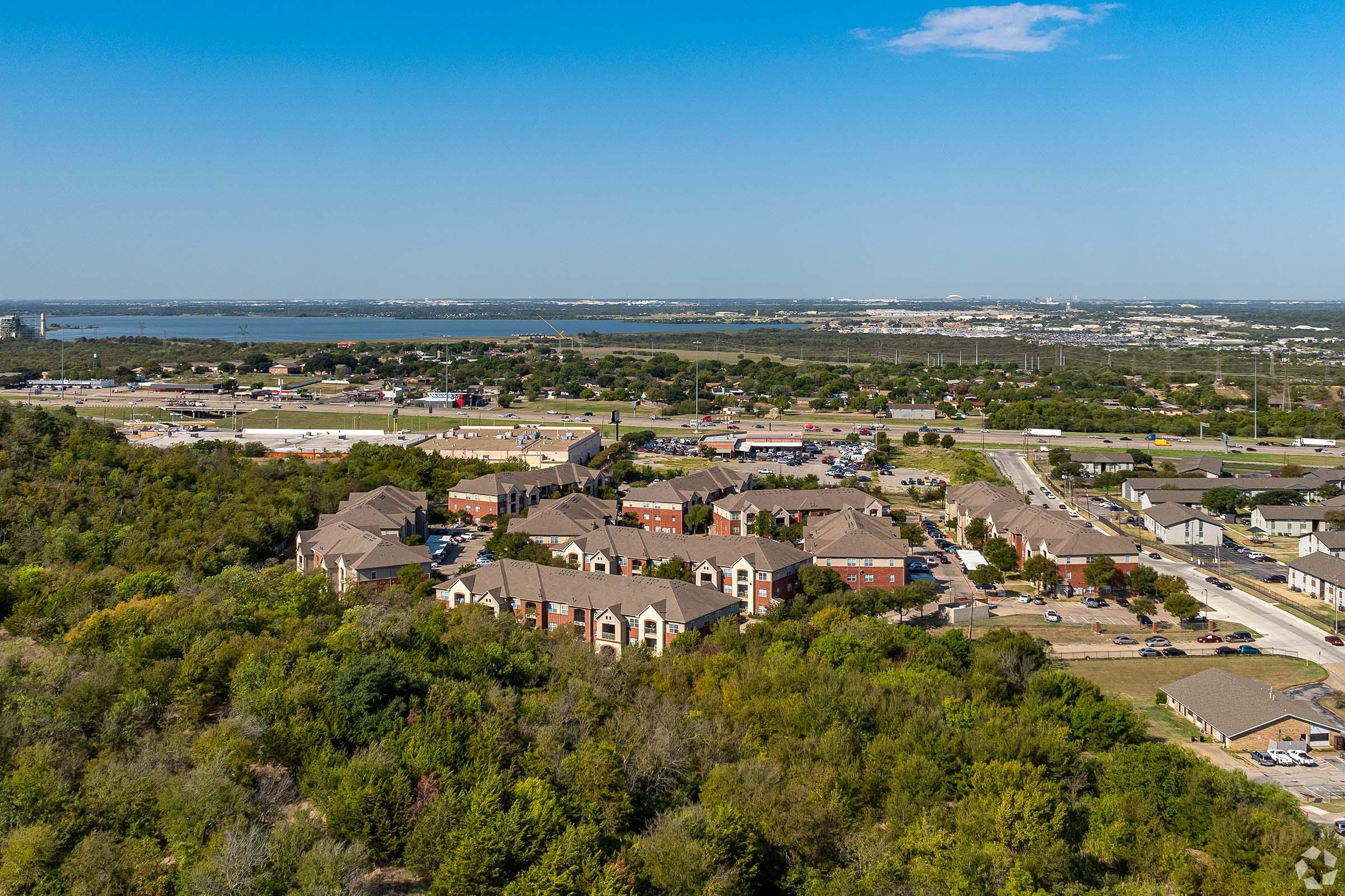 Aerial view of a suburban area featuring residential buildings surrounded by greenery, with parking lots and roads visible. In the background, a waterfront and cityscape can be seen under a clear blue sky. The image captures a blend of urban and natural elements.