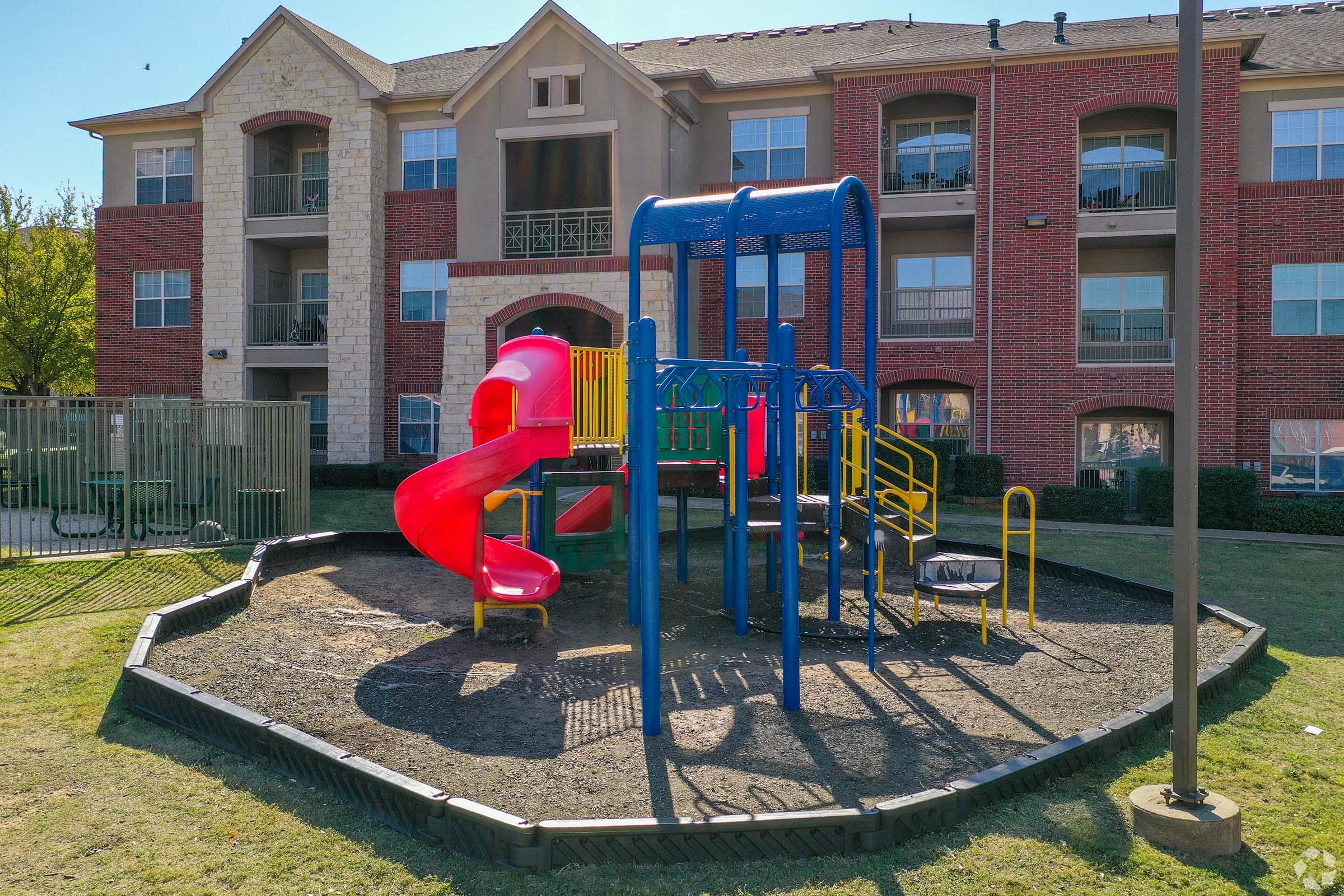 A colorful playground with a red slide, blue climbing structure, and yellow climbing bars, situated on a grassy area in front of a residential apartment complex. The playground is enclosed with a black border and features safety surfacing below the play equipment.