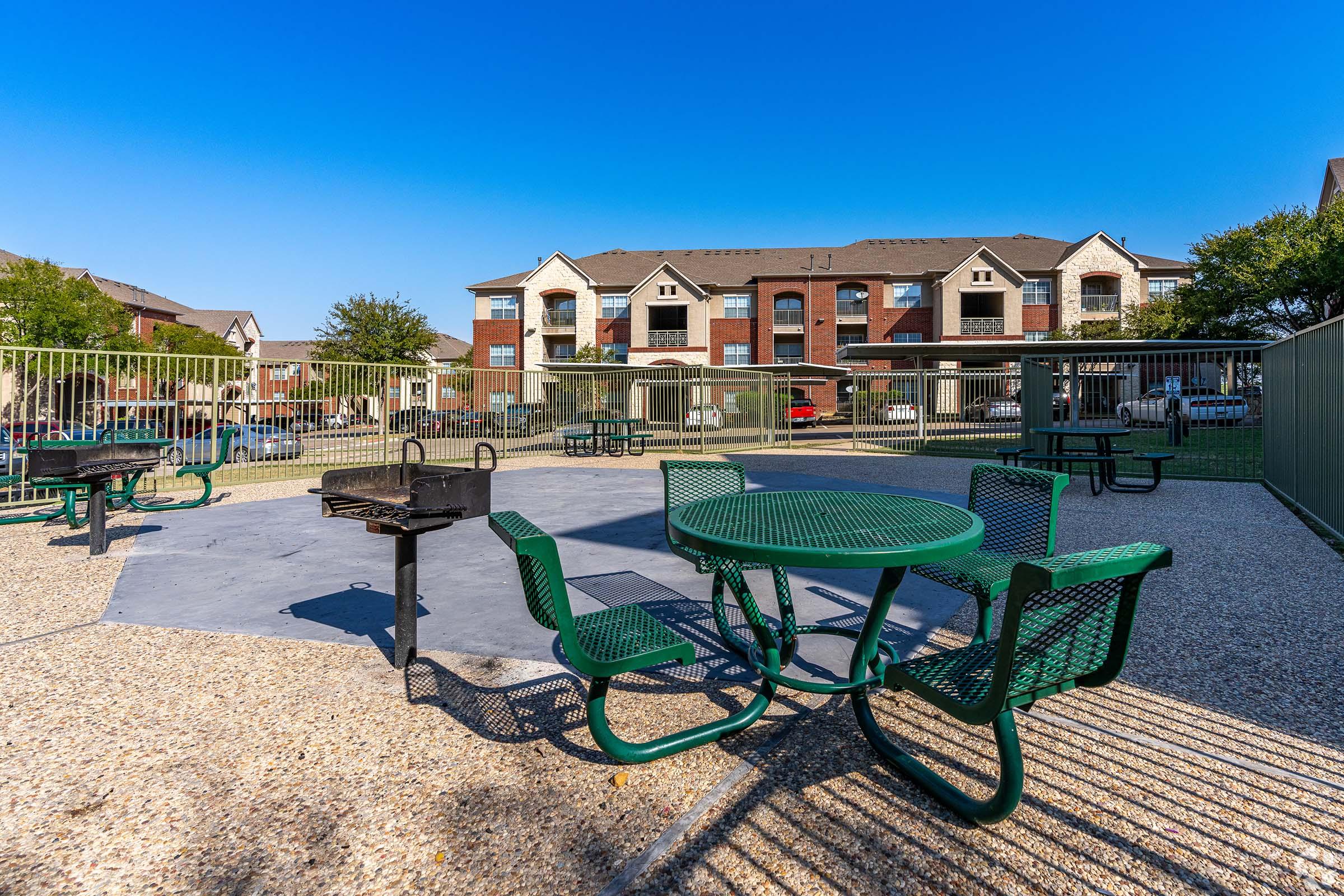 A spacious outdoor BBQ area featuring green picnic tables and a charcoal grill, surrounded by a fence. In the background, residential buildings with multiple apartments are visible under a clear blue sky. The ground is made of gravel, creating a welcoming space for gatherings.