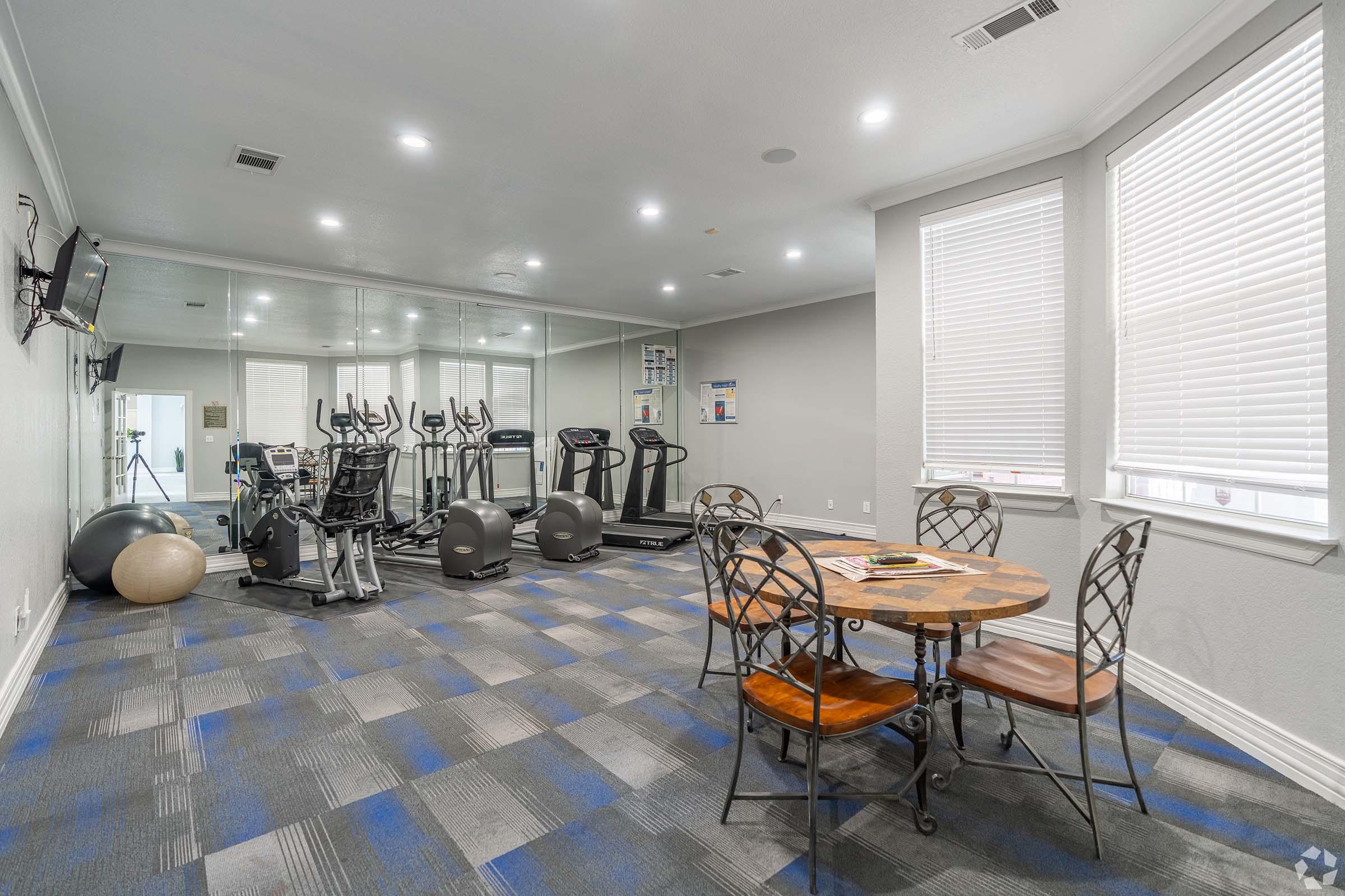 A modern fitness room featuring exercise equipment like treadmills and stationary bikes, a large mirror, and a small dining table with four chairs. The room has bright overhead lighting and large windows with blinds, complemented by a blue and gray carpet.