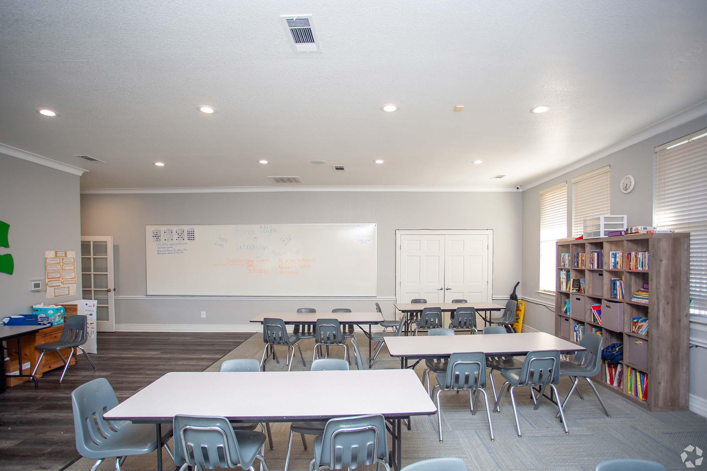 A bright classroom with several tables and chairs arranged for students. A large whiteboard is prominently displayed on the wall, with some writing visible. There is a bookshelf filled with books in one corner, and windows allow natural light to fill the space. The overall atmosphere is inviting and organized.