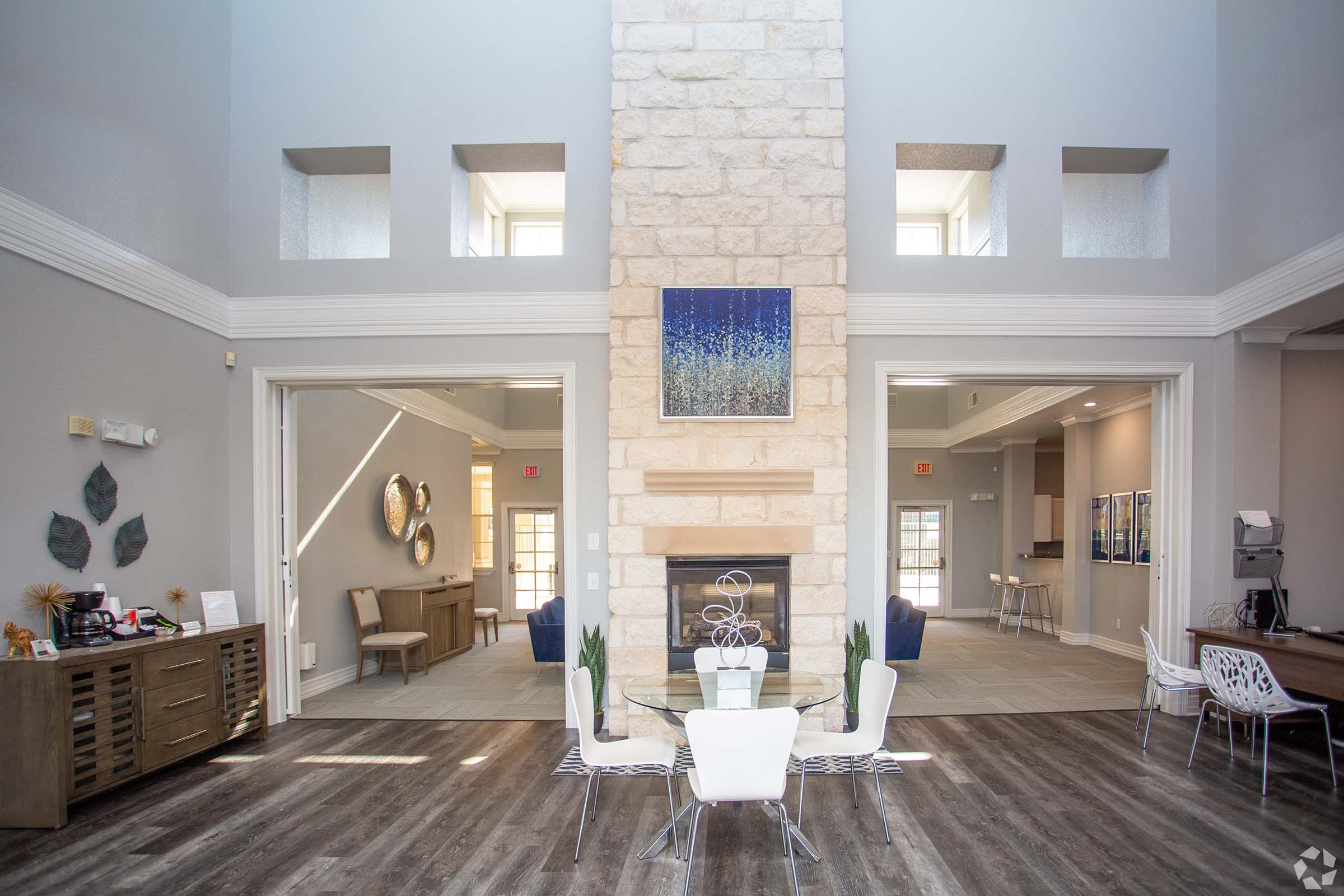 A spacious room featuring a tall stone fireplace with an abstract blue artwork above it. The area has large windows, modern furniture including a round glass dining table with white chairs, and additional seating to the sides. The decor is neutral and inviting, with a contemporary design.