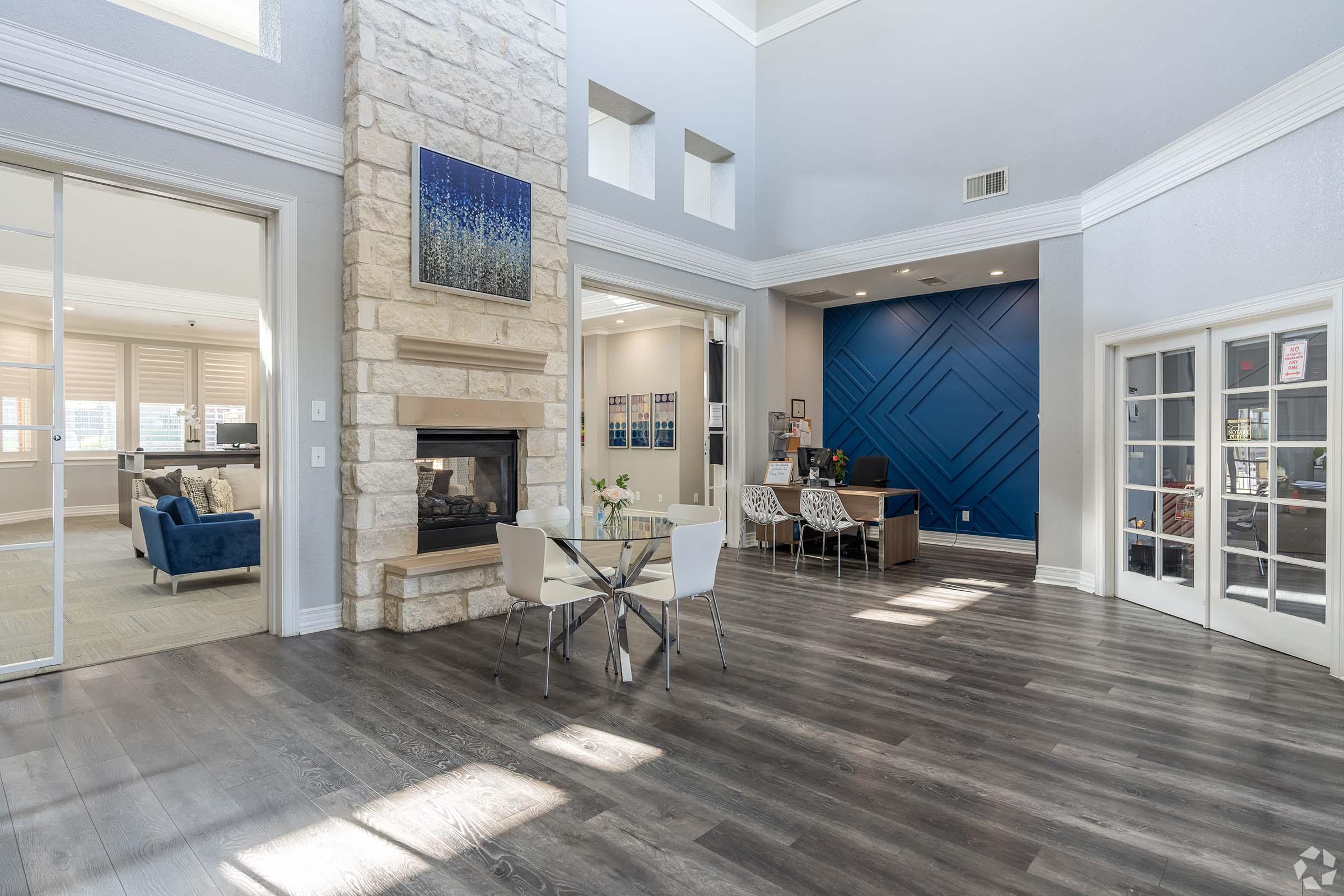 Modern interior space featuring a stone fireplace, light wood floors, and a blue accent wall. A dining table with chairs is positioned near the fireplace, while a glass door leads to another room with comfortable seating. The area has ample natural light from large windows.