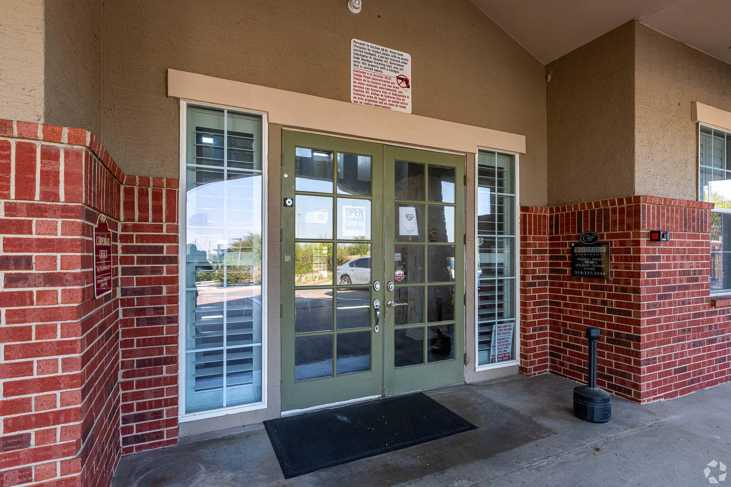Entrance of a building featuring double glass doors framed by green paint, flanked by red brick walls. Above the doors, there is a sign with various rules and regulations. The ground is concrete with a welcome mat in front. The surrounding area appears well-maintained with a glimpse of greenery outside.