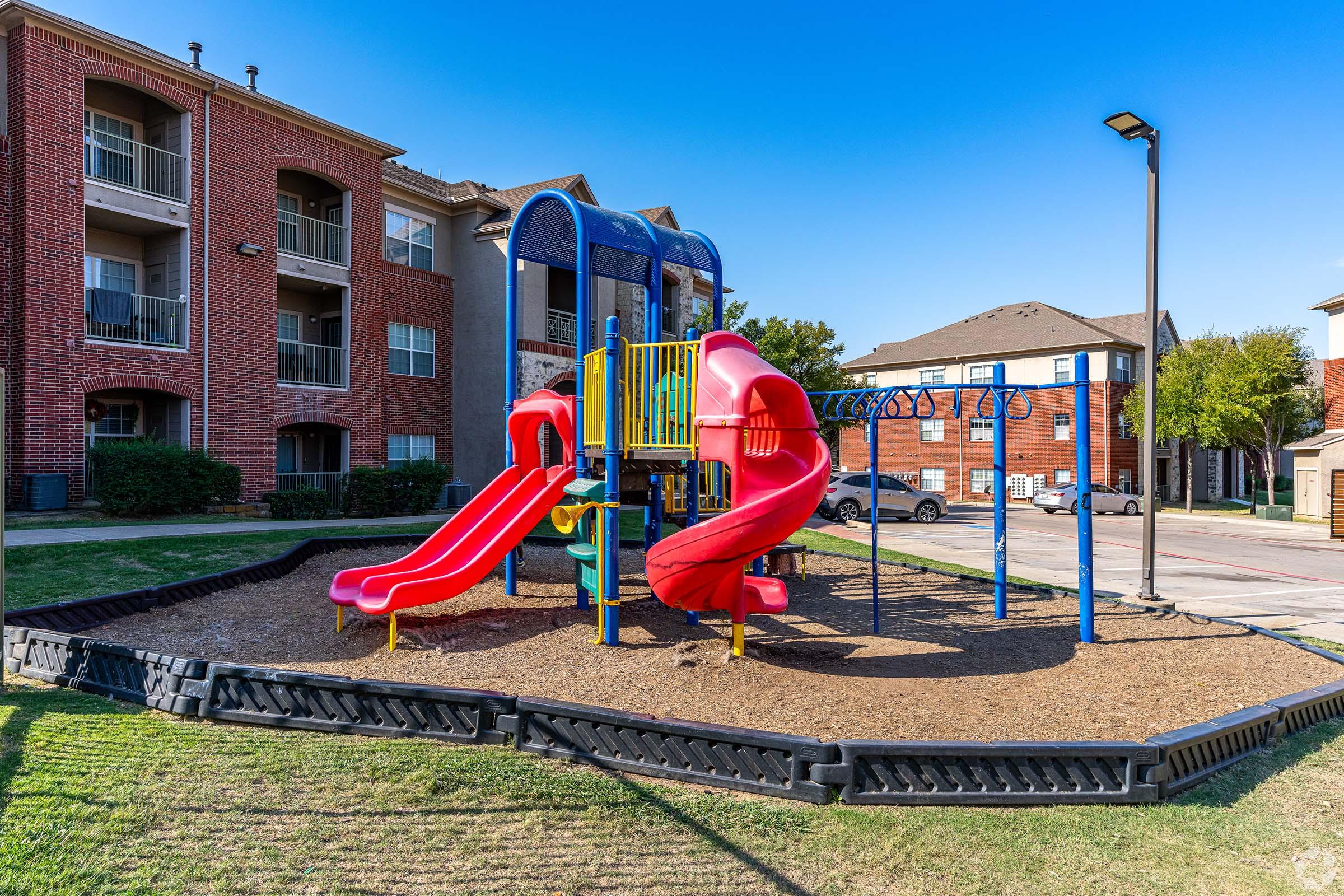 Colorful playground equipment featuring two red slides, climbing structures, and safety features, situated on a grassy area beside residential buildings. The sky is clear with minimal clouds, and the playground is surrounded by a black rubber border for safety.