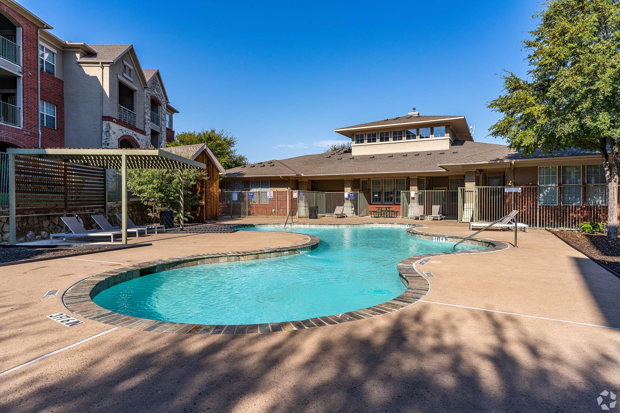 A clear blue swimming pool surrounded by a deck and lounge chairs, with a few trees and a building in the background. The area is well-maintained and features a relaxing atmosphere, perfect for leisure and recreation. The sky is clear and sunny, enhancing the inviting outdoor space.