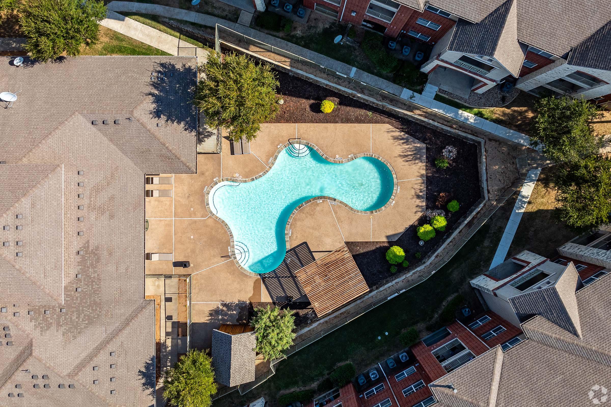 Aerial view of a community pool shaped like a figure eight, surrounded by a patio and landscaped areas. Nearby, residential buildings with brown rooftops are visible, along with green trees and shrubs enhancing the outdoor environment. The pool area features lounge chairs and a shaded structure.