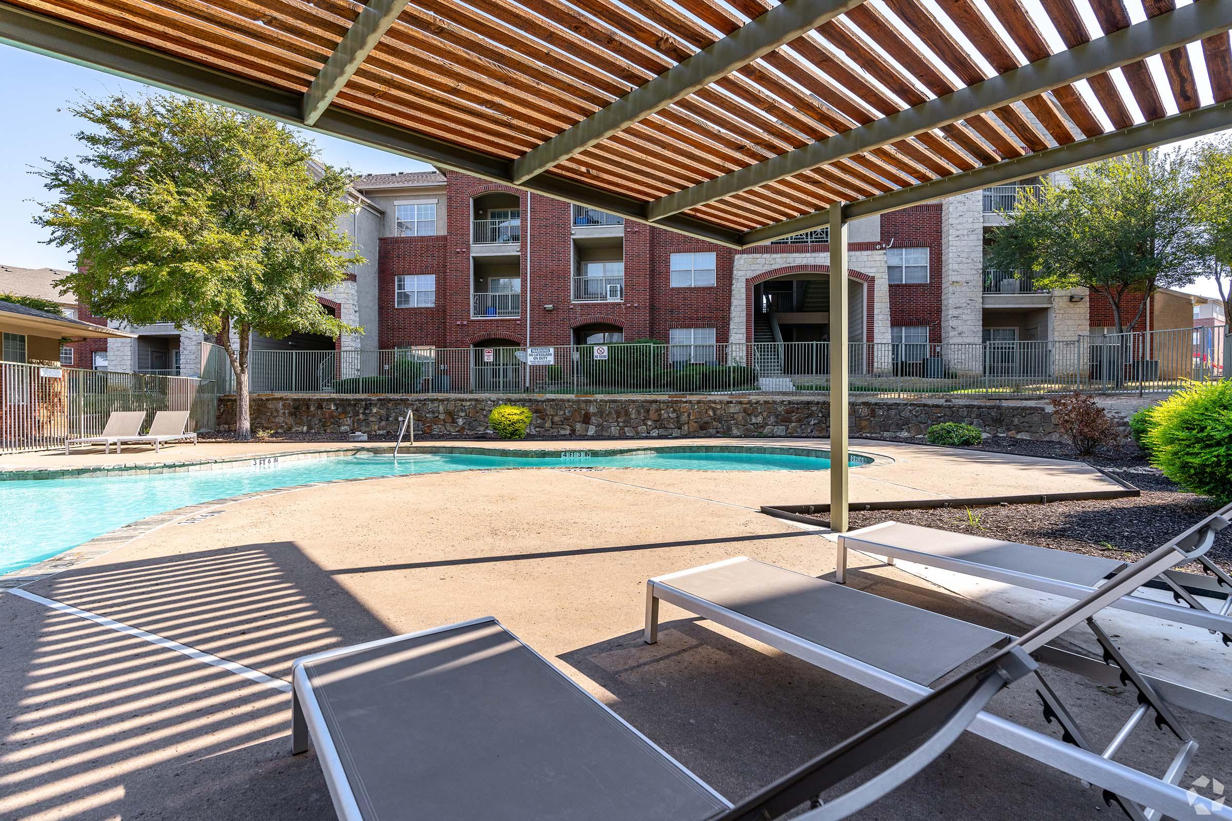 A swimming pool area with lounge chairs under a pergola, surrounded by greenery and residential buildings in the background. The pool is inviting, with clear blue water and a sunny sky overhead, creating a relaxing outdoor atmosphere.