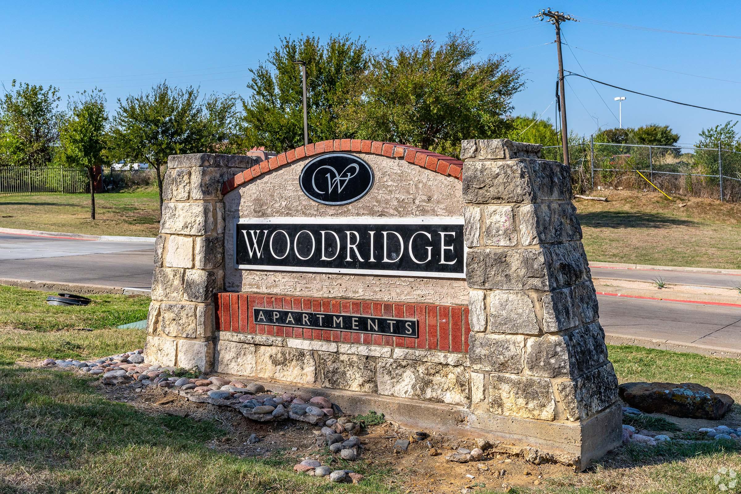 Sign for Woodridge Apartments featuring a stone base and red brick accents. The sign has decorative elements, including the initials "W" and "P" in an emblem. Surrounding greenery and a paved area are visible. The setting appears to be a residential area.