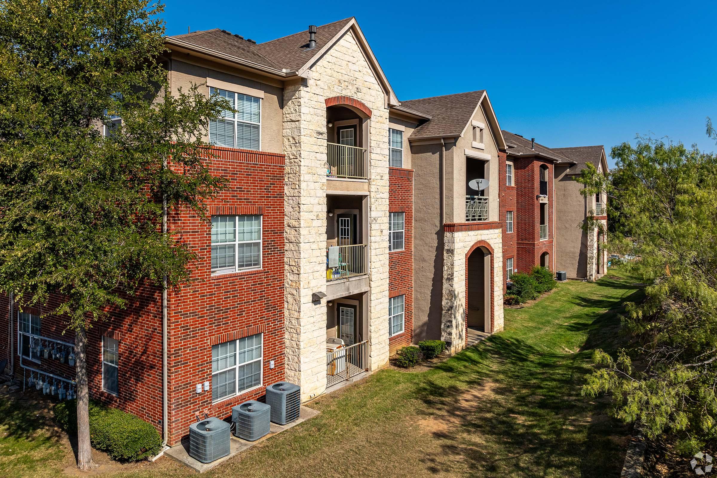 A view of several multi-story apartment buildings featuring a mix of brick and stone facades. Each building has balconies and air conditioning units on the ground level. The surrounding lawn is well-maintained, with trees adding greenery to the scene against a clear blue sky.