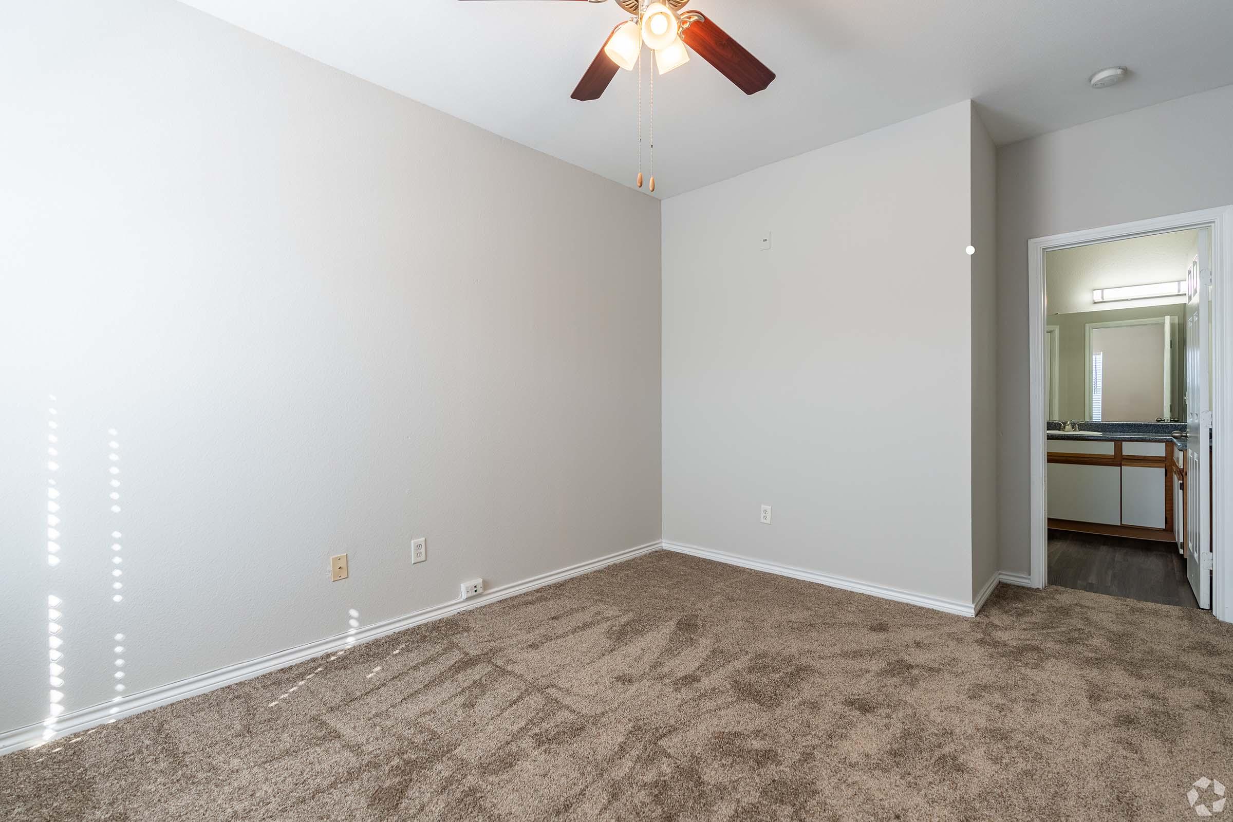 A spacious, light-filled room with beige carpet, a ceiling fan, and neutral-colored walls. The room features a doorway leading to a bathroom with visible fixtures. Sunlight streams through the window, casting soft shadows on the floor. Ideal for a bedroom or study.