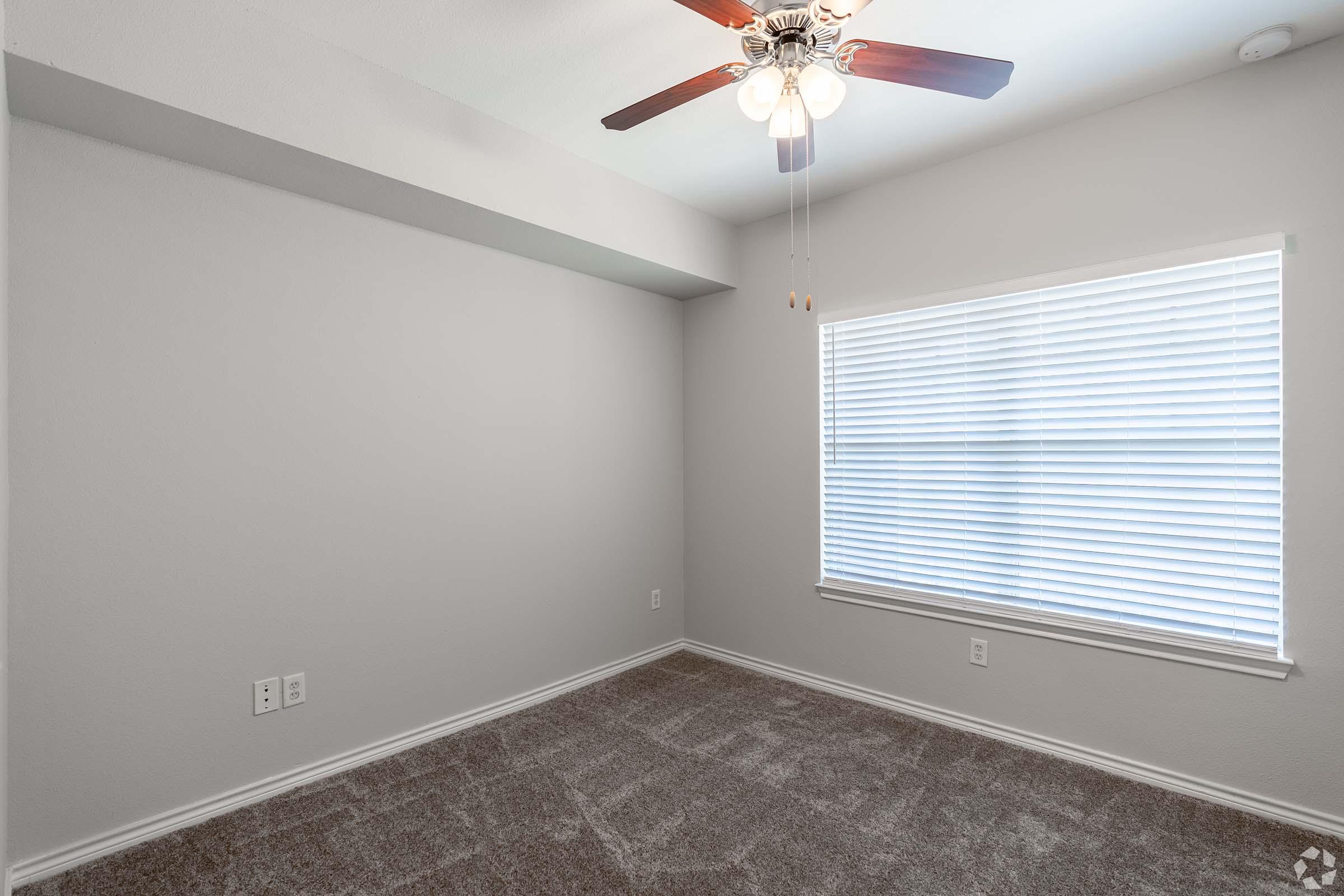 Empty room with light gray walls and carpet. A ceiling fan with wooden blades hangs from the ceiling. There is a window with white blinds allowing natural light to enter, and visible electrical outlets on the wall. The space is clean and ready for furnishing.