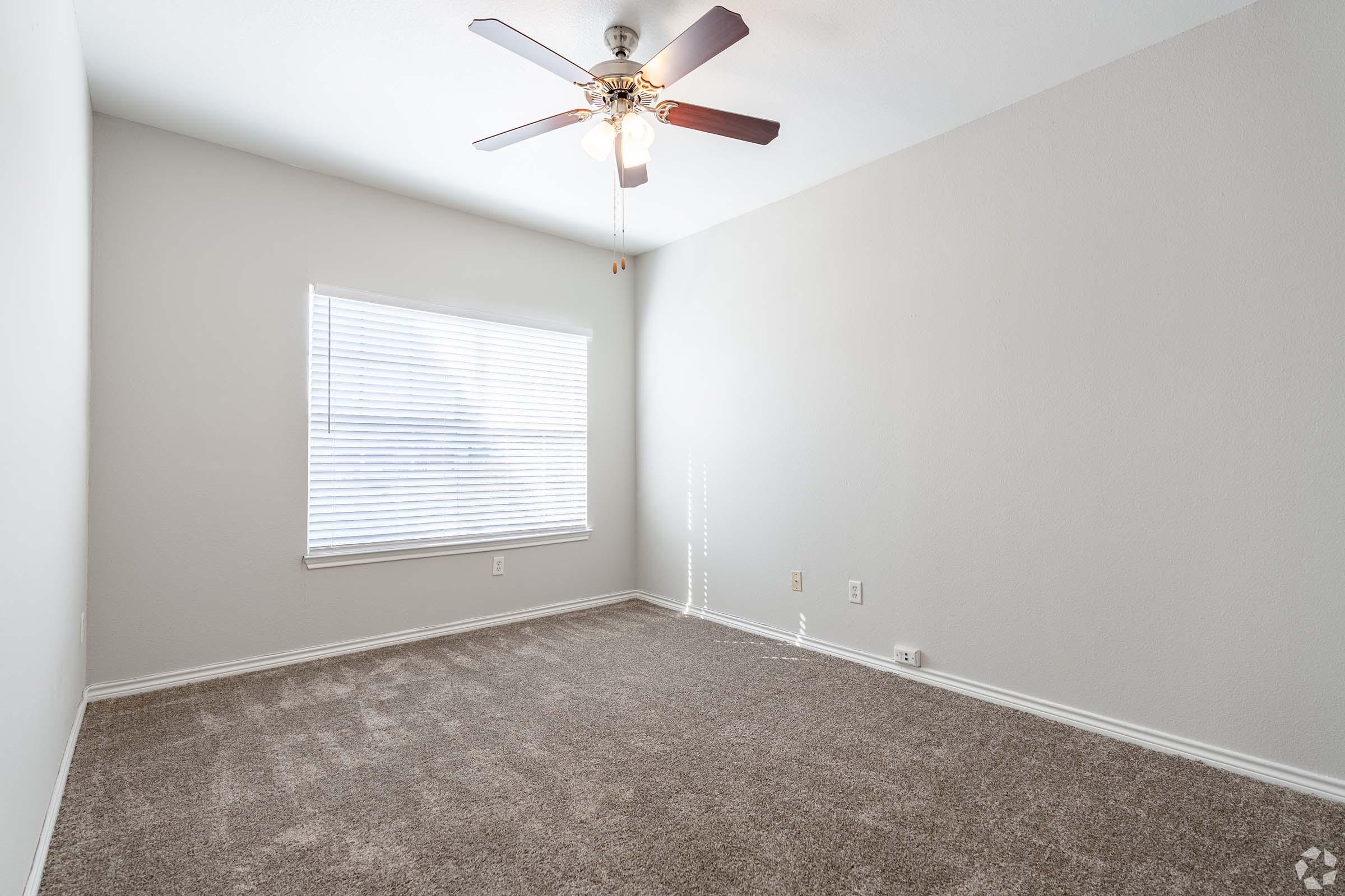 A well-lit, empty room featuring a ceiling fan and a window with blinds. The walls are painted in a light color, and the floor is covered with plush, beige carpet. Sunlight streams in through the window, creating a bright and inviting atmosphere.