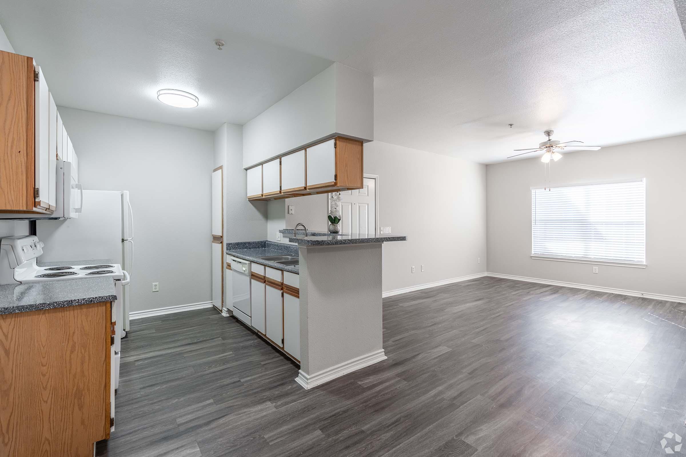 A spacious apartment interior featuring a kitchen with wooden cabinets and a countertop, equipped with an oven, stove, and refrigerator. The living area is bright with natural light from a window and has a ceiling fan. Gray laminate flooring adds a modern touch to the open layout.