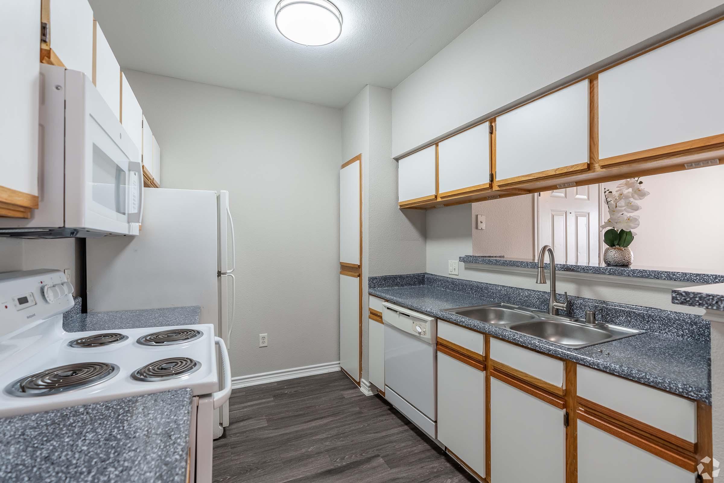 A modern kitchen featuring white appliances, including a stove, microwave, and refrigerator. The countertops are a dark speckled design with a dual sink and a dishwasher. Cabinets are a light wood color, and the walls are painted in a neutral tone. Ample natural light fills the space from an overhead fixture.