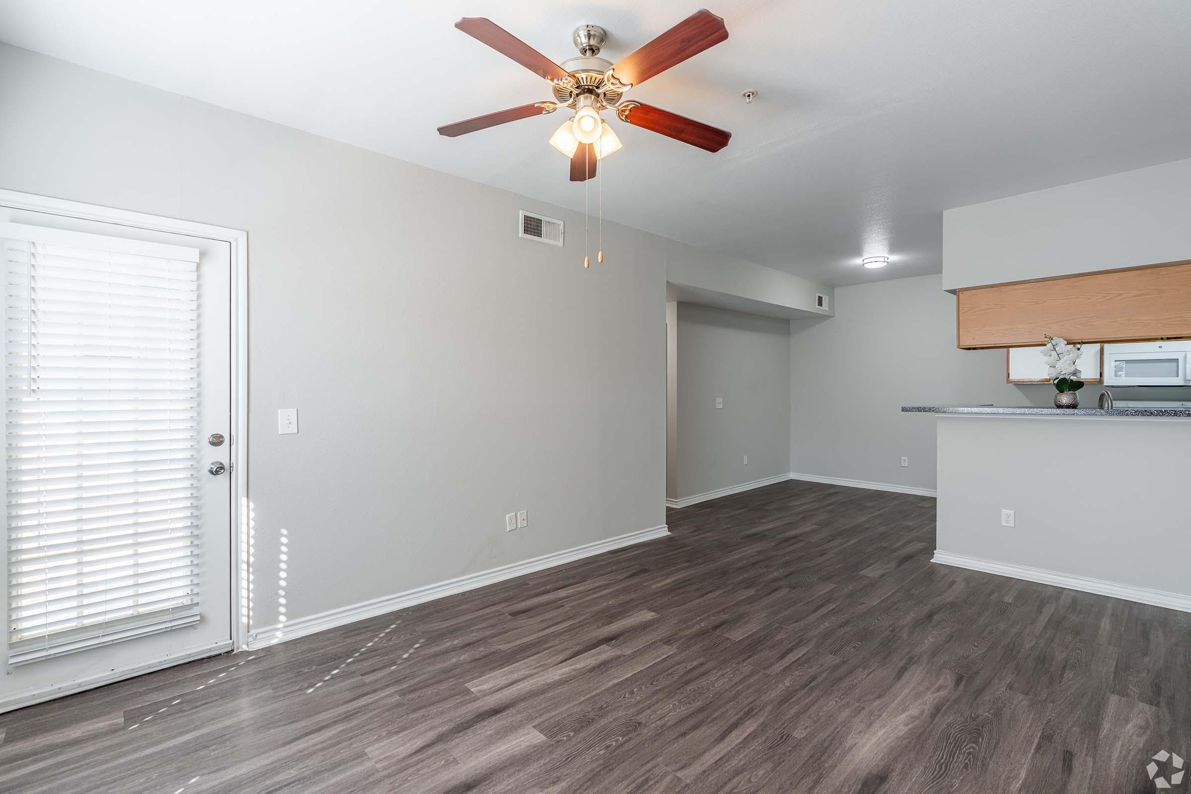 A well-lit, spacious living area with light gray walls and modern laminate flooring. It features a ceiling fan, a light wood kitchen counter visible in the background, and a glass door leading outside. The design is clean and contemporary, making it suitable for various interior styles.