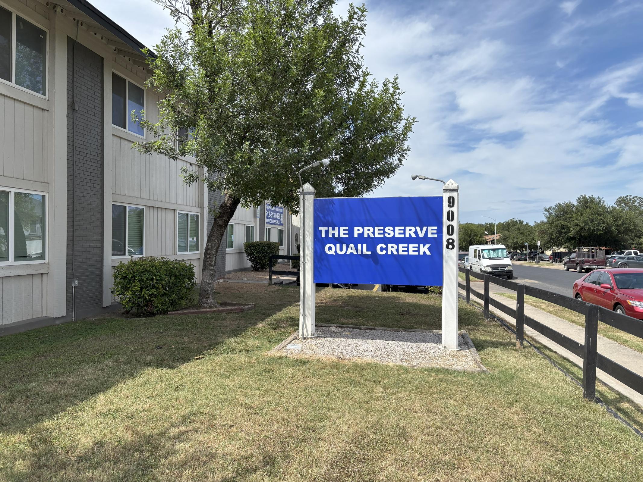Sign for "The Preserve Quail Creek" displayed prominently in front of a building, with a blue background and white text. Surrounding area features grass, a tree, and nearby parking. Sky is partly cloudy, indicating a bright day. The sign includes a street number, 9008.