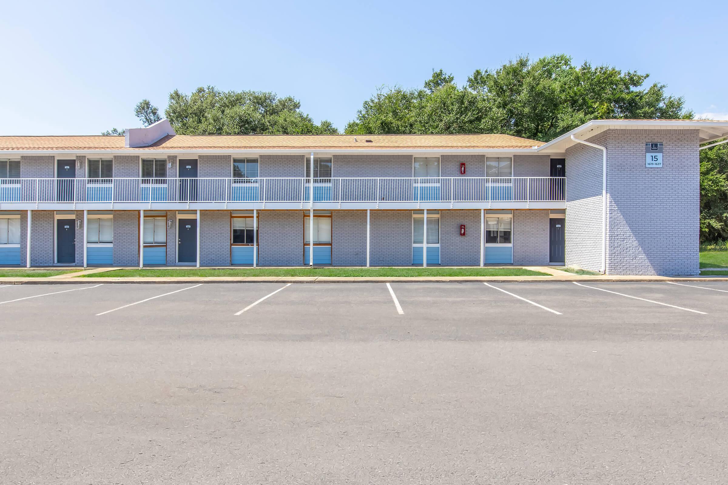 A two-story motel building with multiple rooms, each featuring a balcony. The exterior is light gray with a tiled roof. In front, there is a spacious parking lot with several empty spaces, and trees line the background under a clear blue sky.
