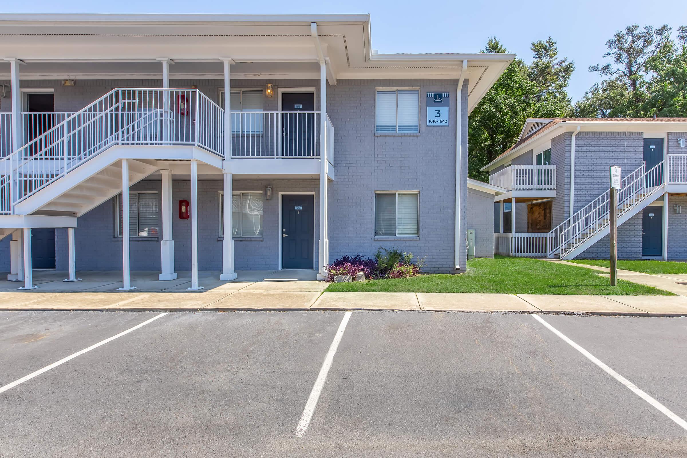 A view of two multi-unit residential buildings with a gray exterior. One building features a set of stairs leading to the second floor, a door labeled "3," and well-maintained landscaping. Parking spaces are visible in the foreground, with a clear blue sky overhead.