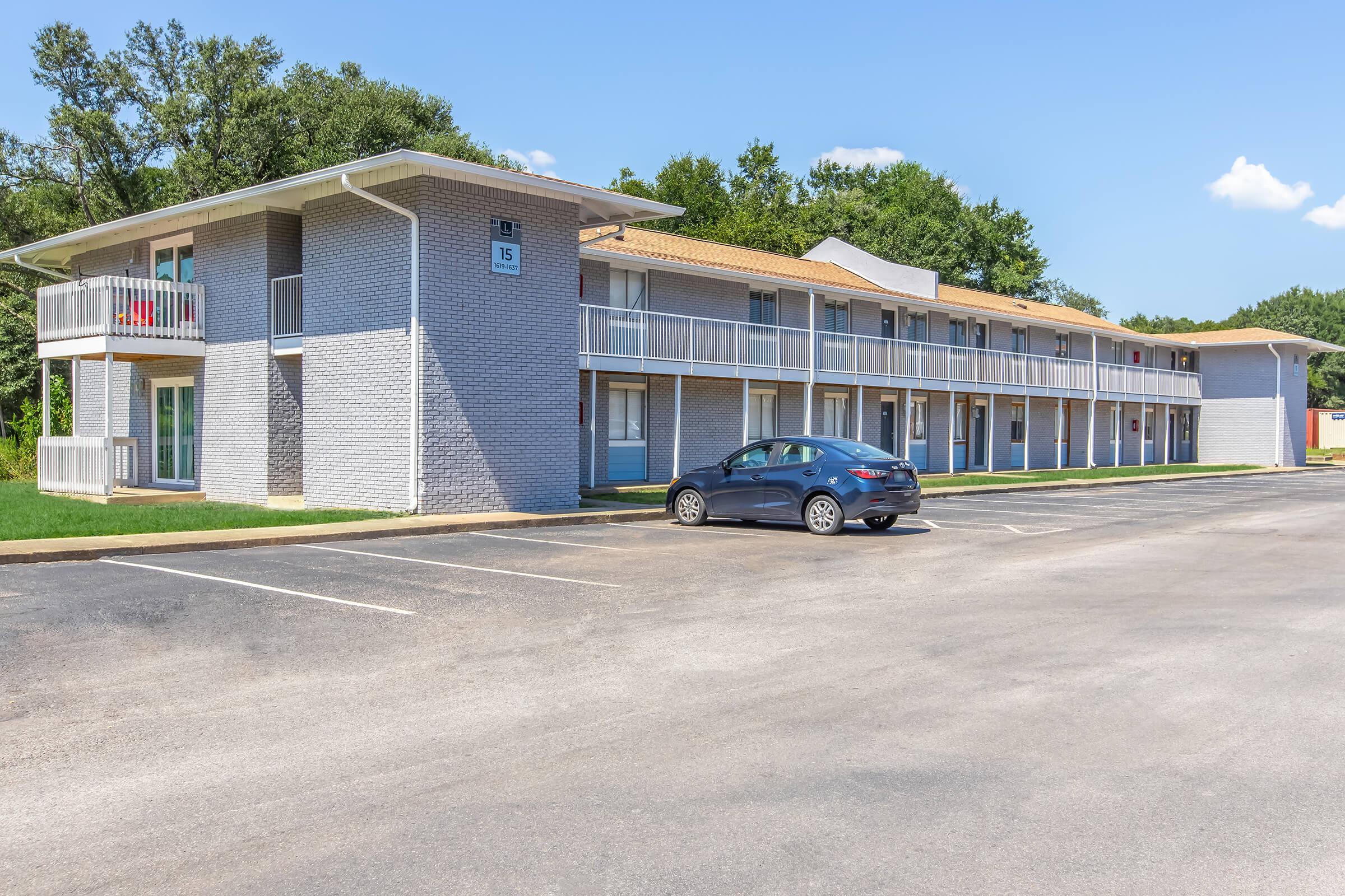 A two-story, modern motel building with a light gray exterior and attached balconies. Several parked cars in the foreground, with a clear blue sky overhead and green trees in the background. The setting appears well-maintained and inviting.