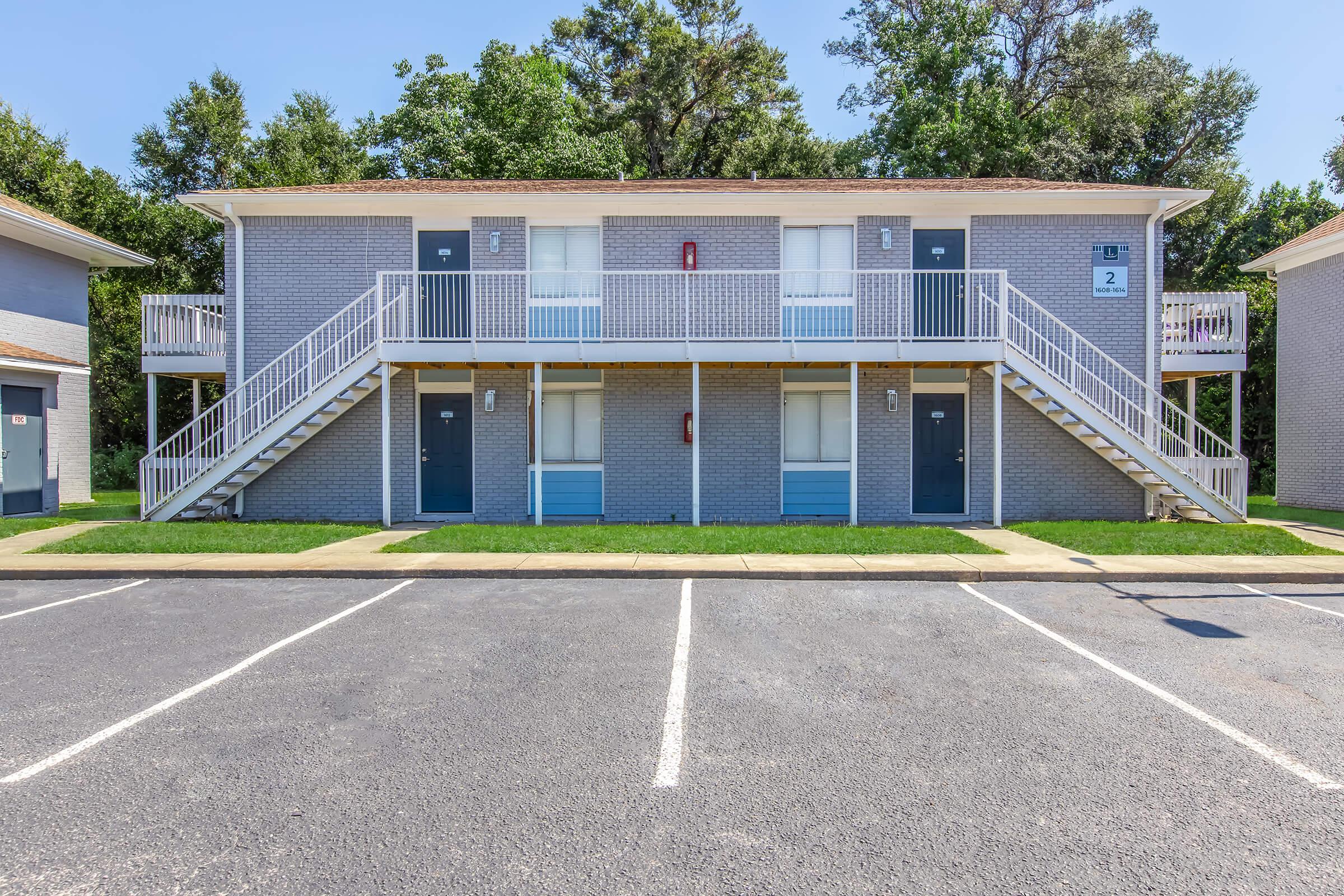 A two-story apartment building with a gray exterior, featuring blue doors and white railings. Two sets of stairs lead to the upper level. The parking lot in front has marked spaces, and there are trees in the background under a clear blue sky.