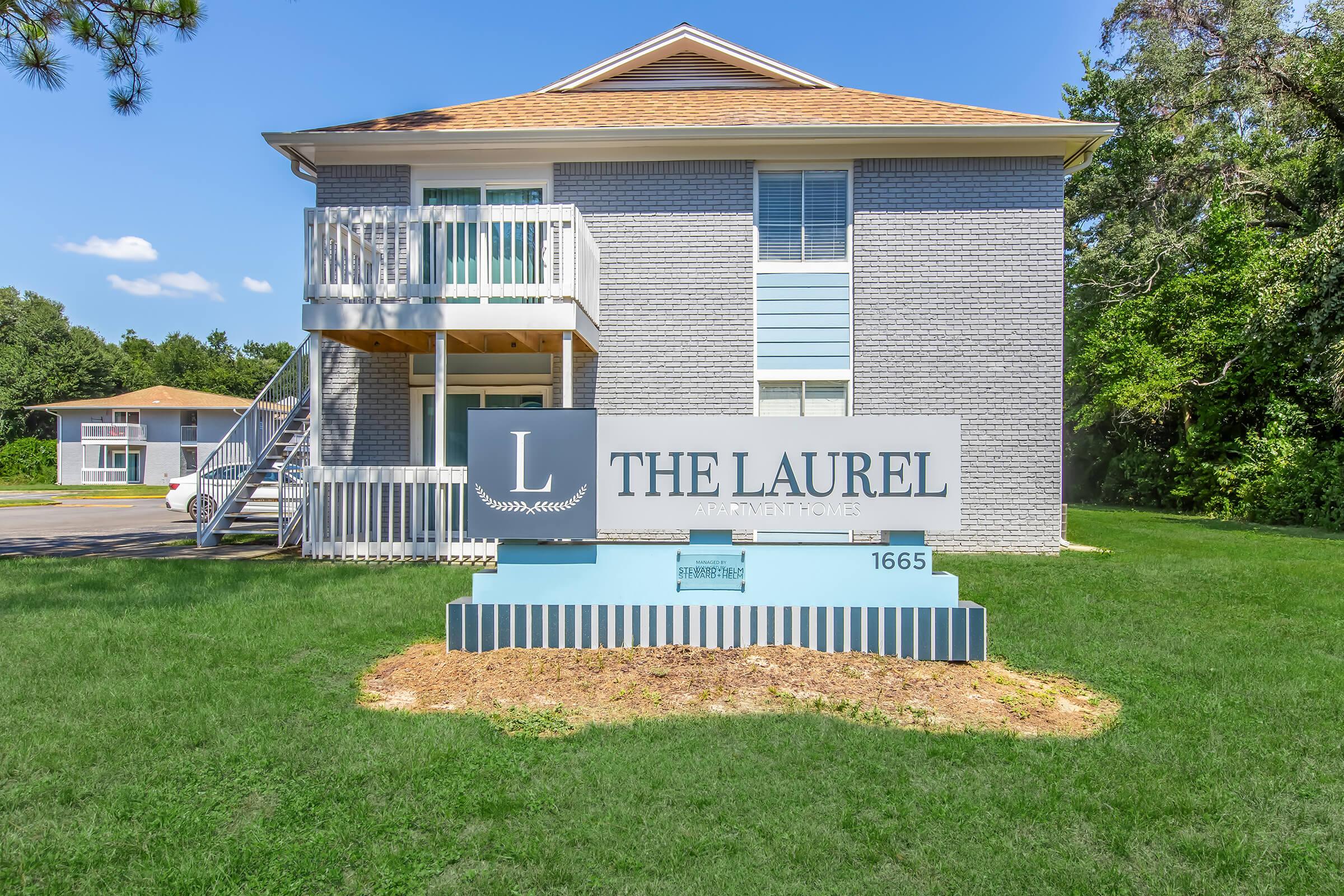 A two-story gray residential building with white trim and a wooden staircase leads to the second floor. A large sign in front displays "The Laurel" along with the address "1665," set in a grassy area with trees in the background.