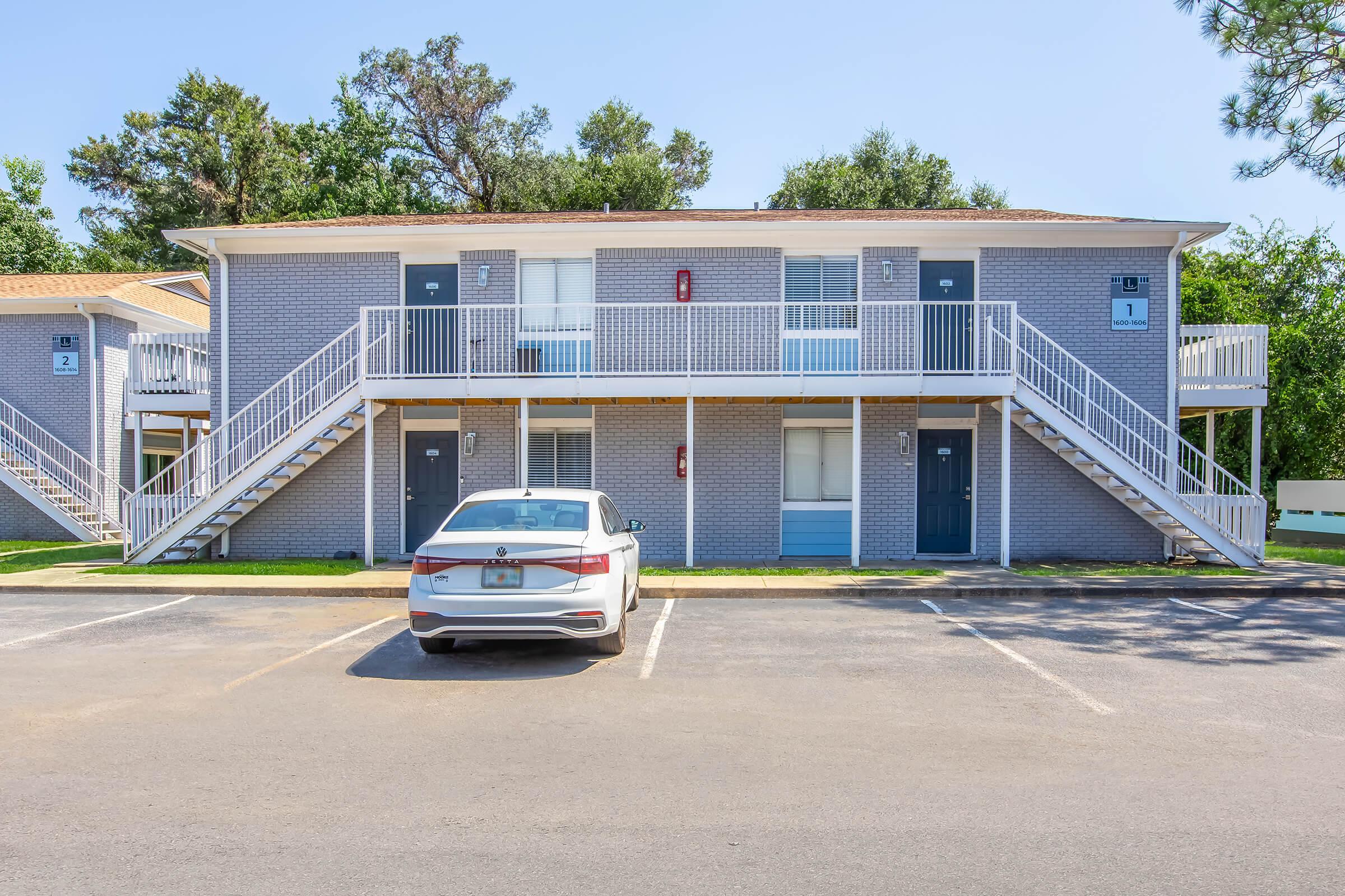 A two-story brick apartment building with blue doors and white railings. There are two sets of stairs leading to the upper level. A white car is parked in the foreground on an asphalt lot, with designated parking spaces visible. Surrounding greenery and trees provide a natural backdrop.