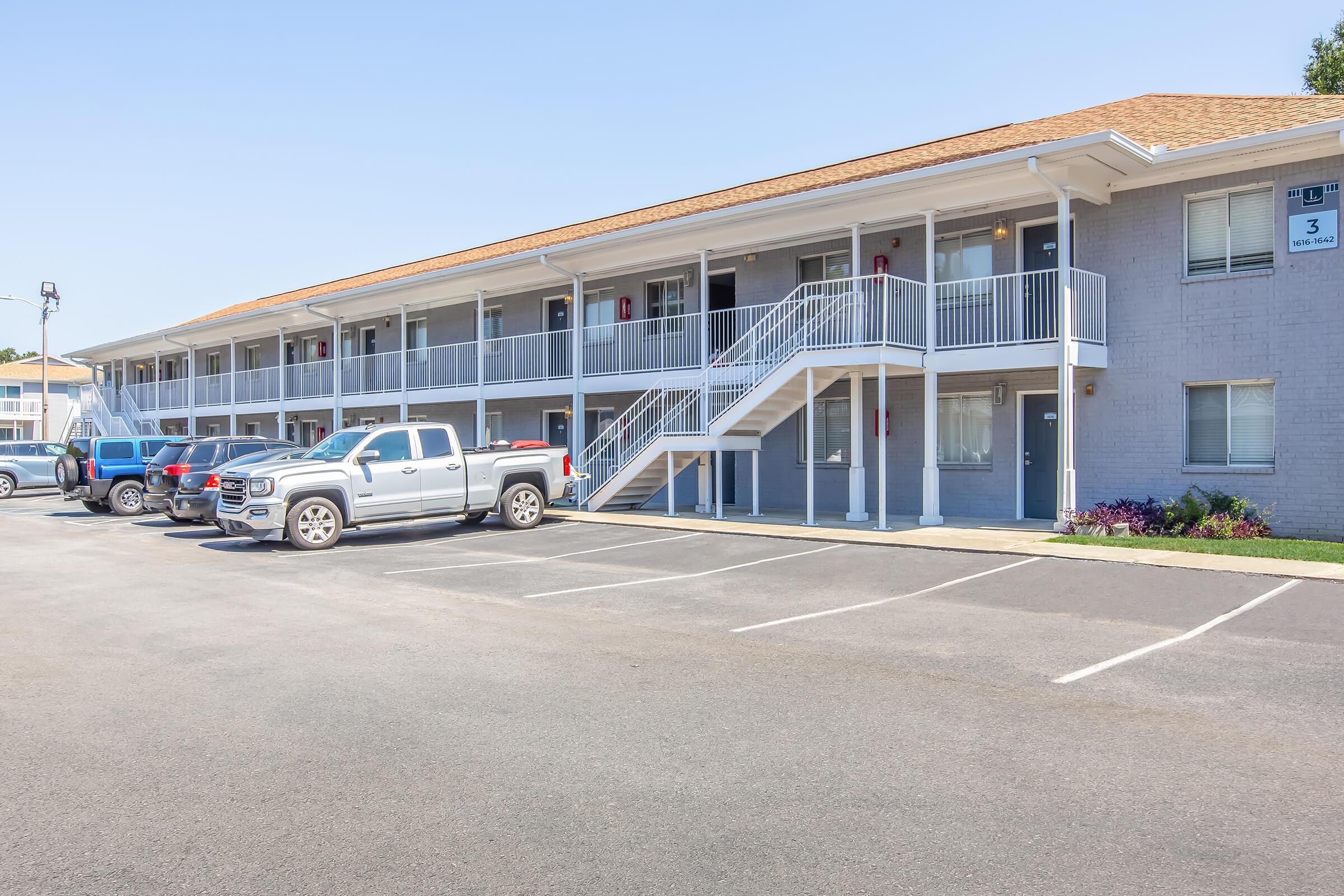 A view of a two-story motel building with a light gray facade. Several parked vehicles are in the foreground, including a silver pickup truck. The entrance features a staircase leading to the second floor, with rooms visible on each side. The parking lot is well-maintained, and there is a clear blue sky above.