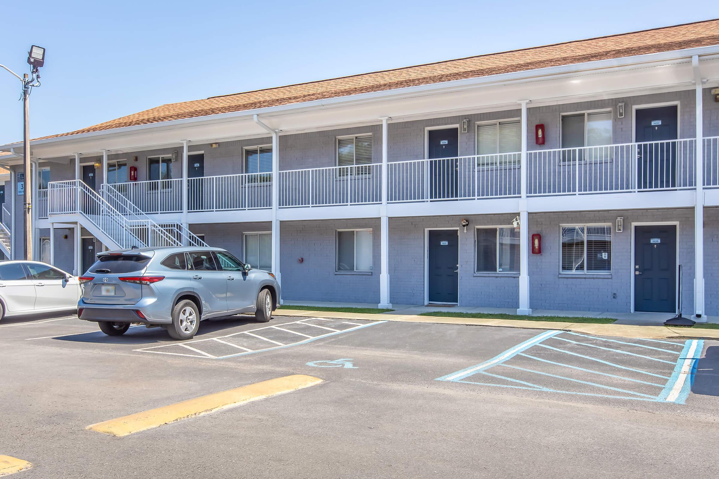 A low-rise motel with two levels of rooms, featuring gray exterior walls and white railings. A parked gray SUV is visible in the foreground, along with designated parking spaces. The entrance to the rooms is visible, with each room having a door and a window. Clear blue skies are in the background.