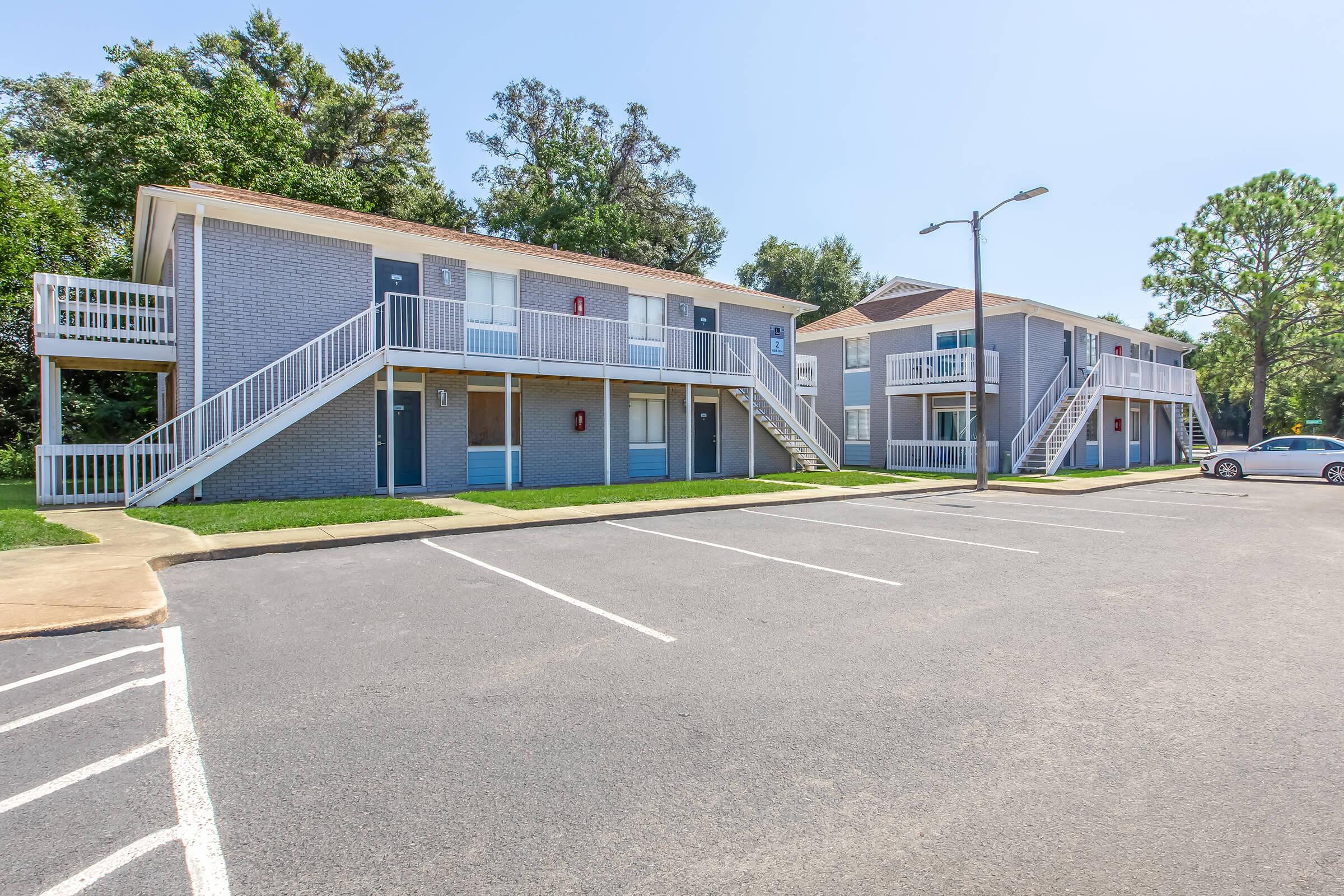 Exterior view of a two-story apartment complex with a light gray brick facade. The building features multiple entrances with staircases leading to the second floor. A neatly paved parking area is in front, with a few parking spaces visible and a light pole. Lush greenery surrounds the property on a clear, sunny day.